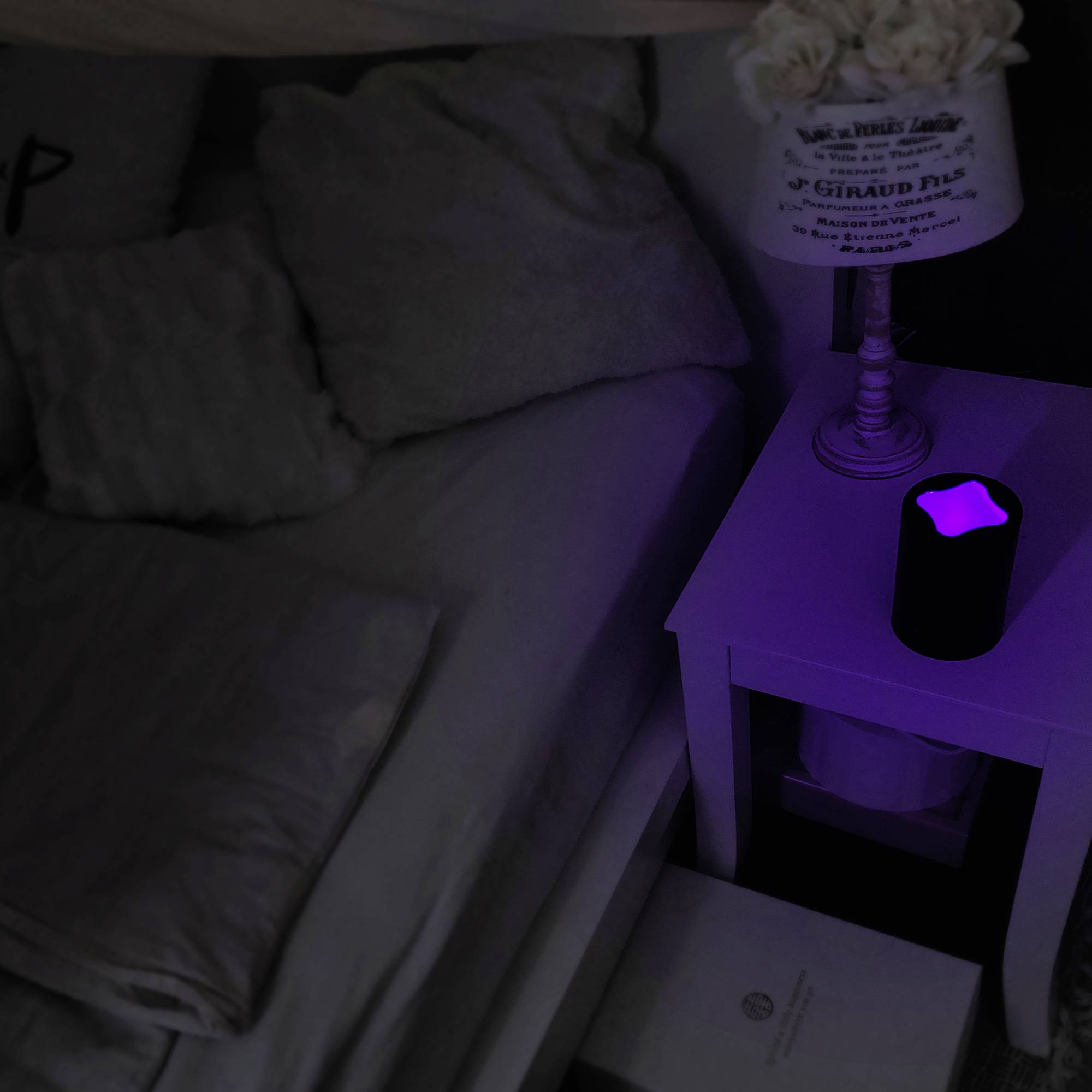 Ein Nachttisch mit einer kleinen, leuchtenden Lampe in dunklem Raum neben einem Bett mit hellem Bettzeug.