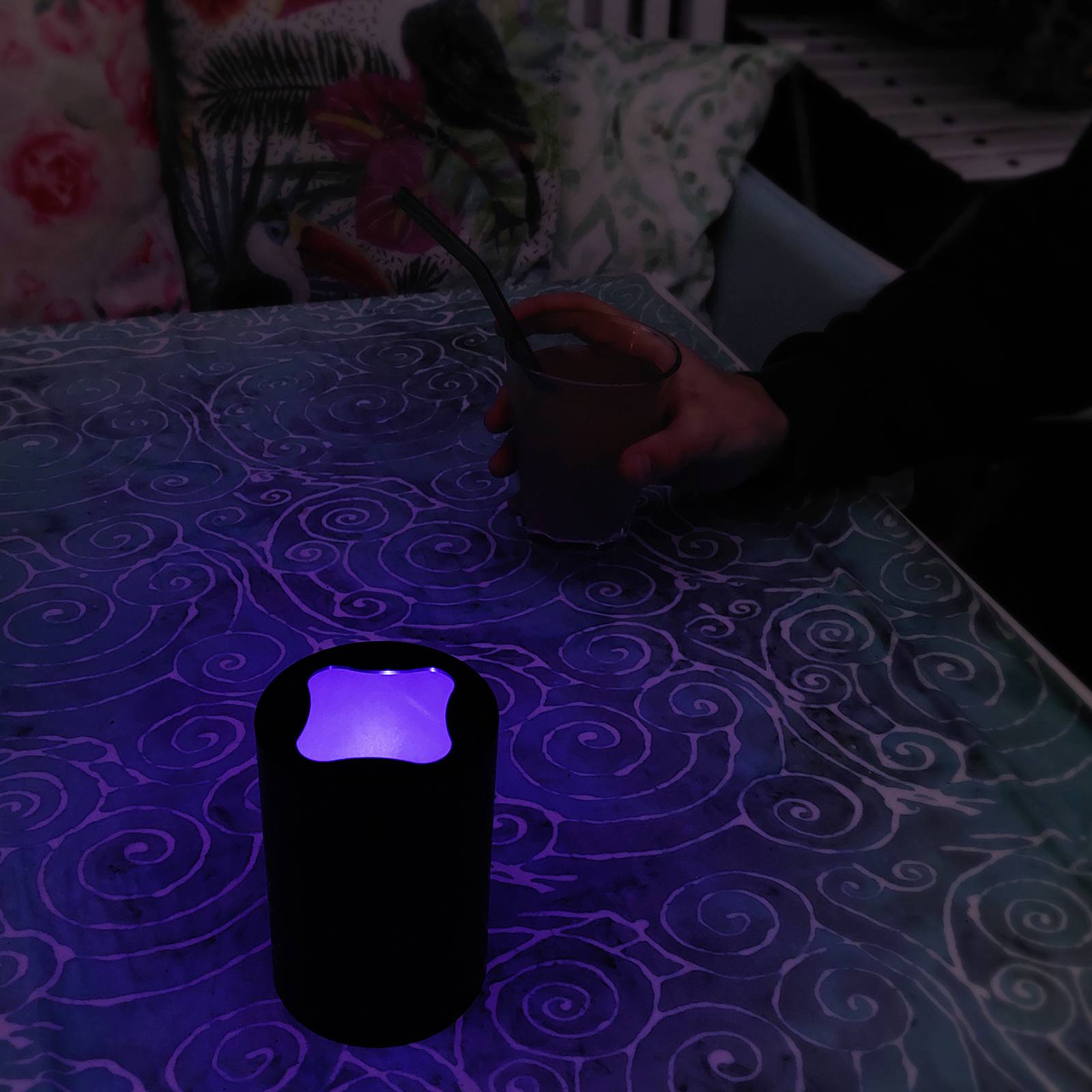 Ein Getränk steht in einem beleuchteten Becher auf einem Tisch mit floralem Muster. Jemand hält einen weiteren Becher. Atmosphärische Szene.