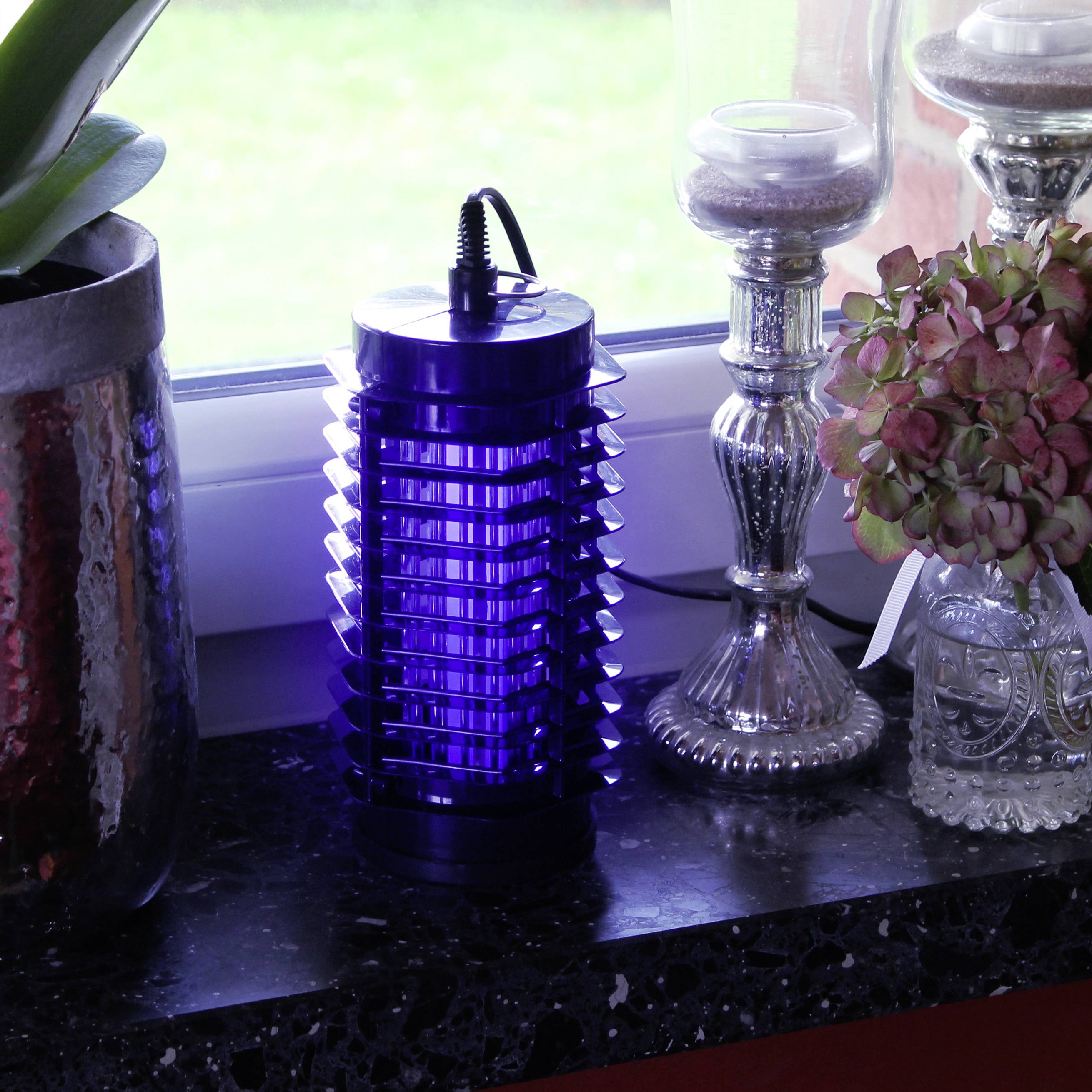 Eine blaue Insektenlampe steht auf einer Fensterbank, flankiert von Pflanzen und Kerzenhaltern. Draußen ist grüne Vegetation sichtbar.