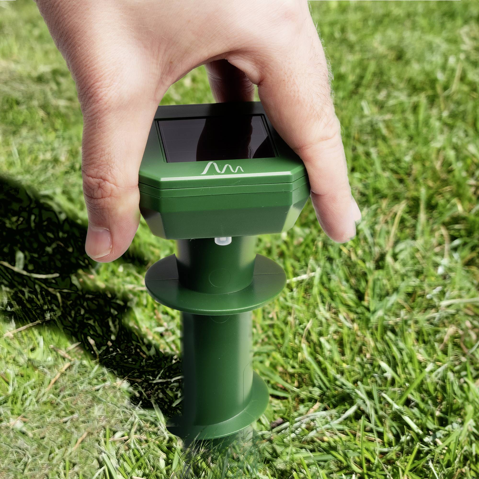 Eine Hand hält ein grünes, solarbetriebenes Vertreibungsgerät für kleine Tiere, das auf einer Wiese platziert ist.