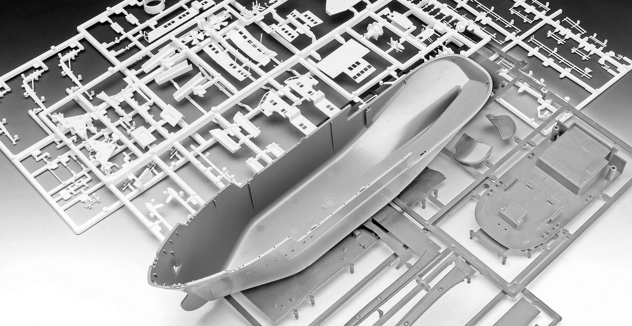 Ein graues Modellbausschiff in Einzelteilen liegt auf einer Bauplatte. Es zeigt den Rumpf und andere Komponenten, bereit zum Zusammenbau.