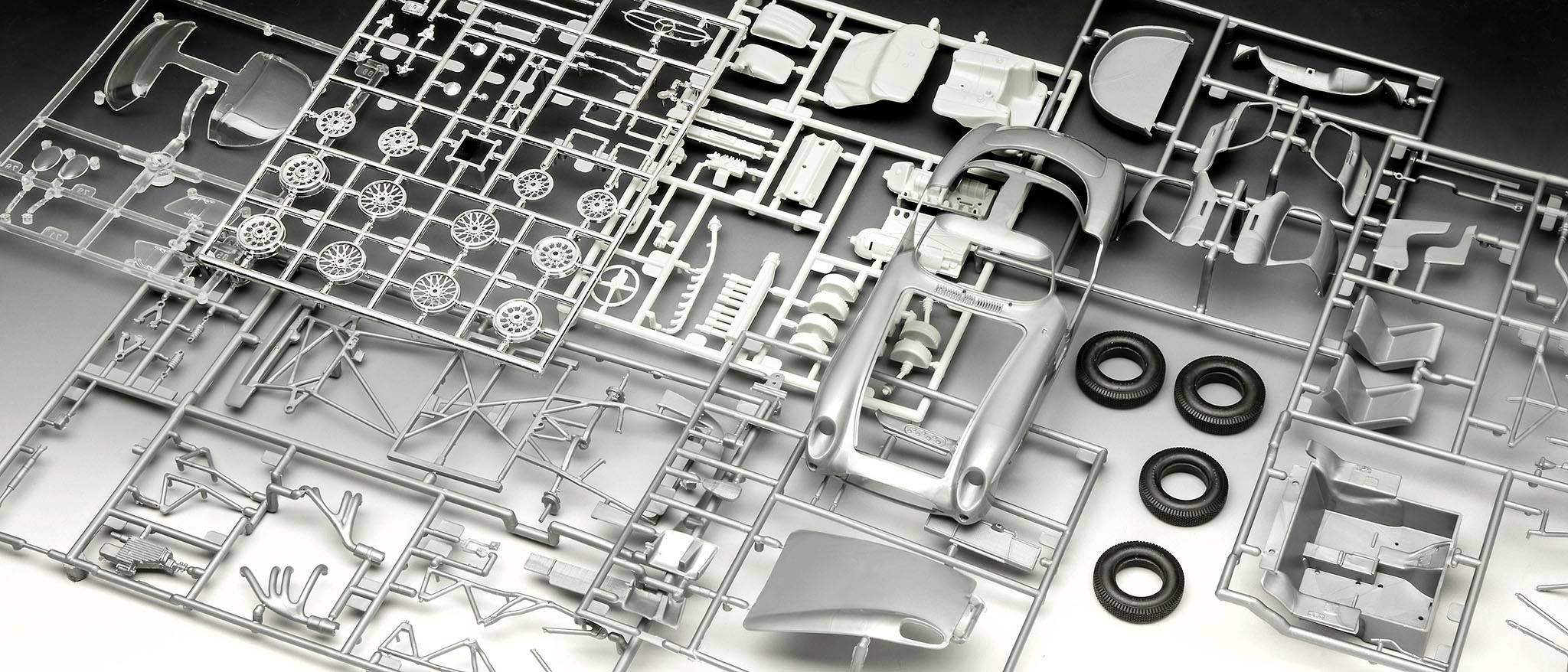 Plastikmodellbausatz auf einem Tisch mit verschiedenen Bauteilen und vier schwarzen Reifen. Ideal für Modellbauprojekte.