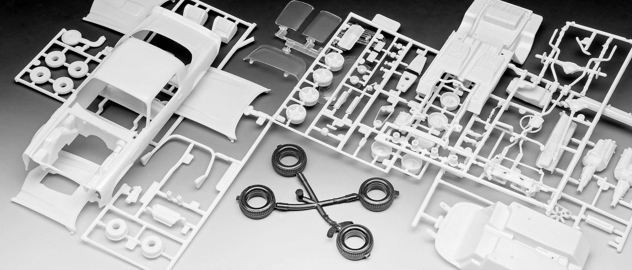 Bausatz eines Modellautos mit verschiedenen weißen Kunststoffteilen und schwarzen Reifen auf grauer Fläche, bereit zum Zusammenbau.
