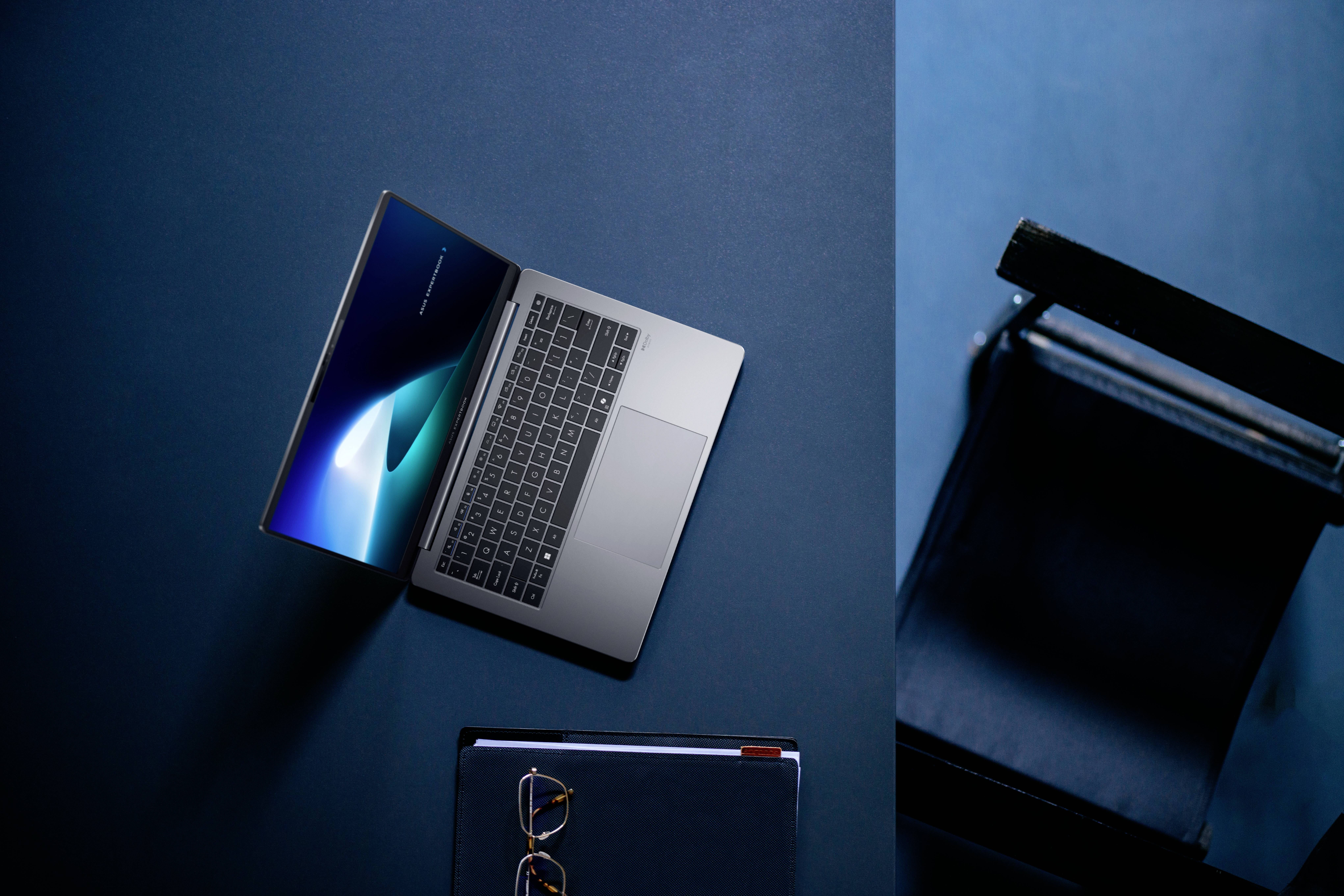 Ein Laptop auf einer blauen Oberfläche neben einem Stuhl und einem geschlossenen Notizbuch mit Brille, der eine Arbeitsplatzeinrichtung vermittelt.