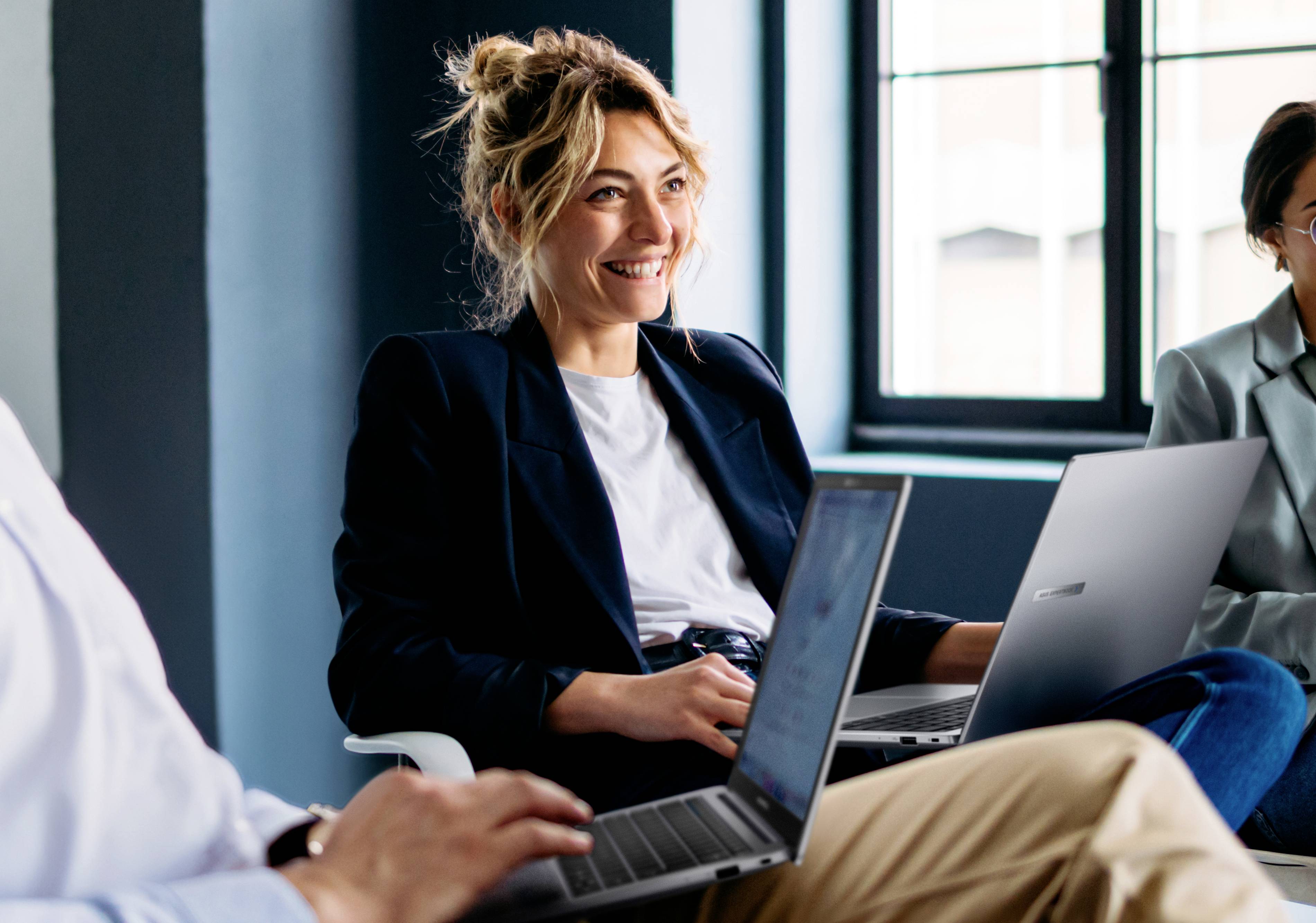 Eine lächelnde Person sitzt in einem modernen Büro mit Laptops und ist in eine kollaborative Arbeitsumgebung eingebunden.