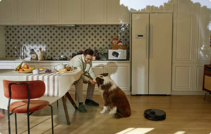 Eine Person sitzt in einer Küche am Tisch und streichelt einen Hund. Im Raum befinden sich ein gedeckter Tisch, ein Kühlschrank und ein Saugroboter.