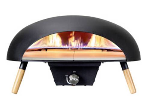 Ein tragbarer Pizzaofen im Freien, der Pizza mit sichtbaren Flammen im Ofenraum backt. Der Himmel ist blau, grüne Wiese im Hintergrund.