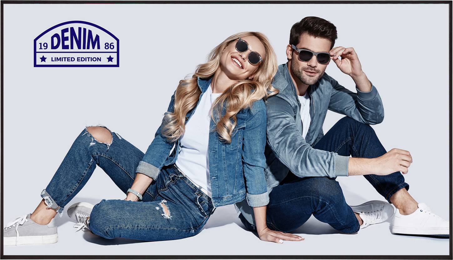 Zwei Personen in Jeansjacken und -hosen sitzen Rücken an Rücken, lächeln und tragen Sonnenbrillen. Ein Logo mit der Aufschrift 