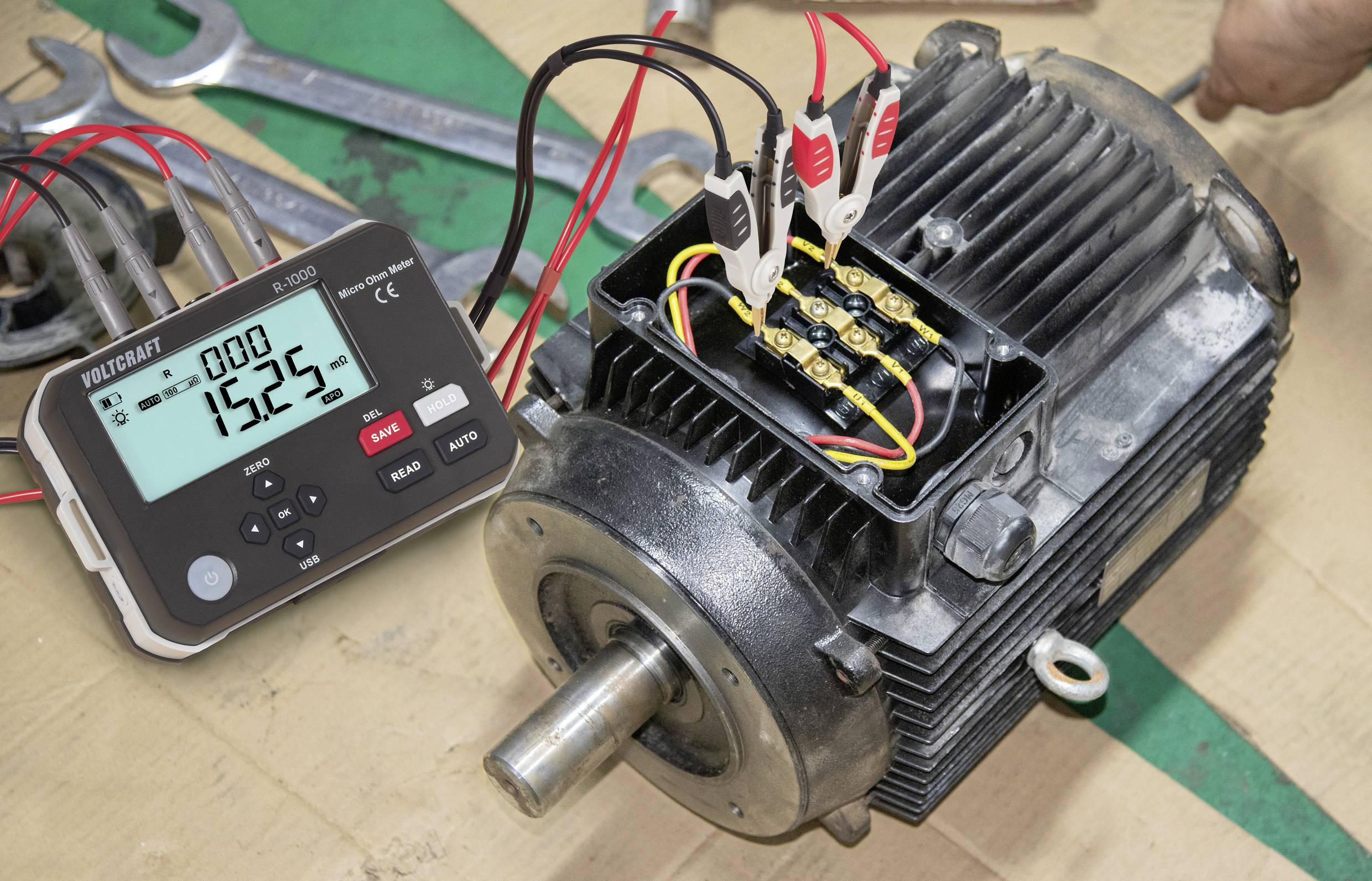 Elektromotor mit Kabeln, die an einen Multifunktions-Kalibrator angeschlossen sind, der '1525' anzeigt. Schraubschlüssel im Hintergrund deuten auf Wartungsarbeiten hin.