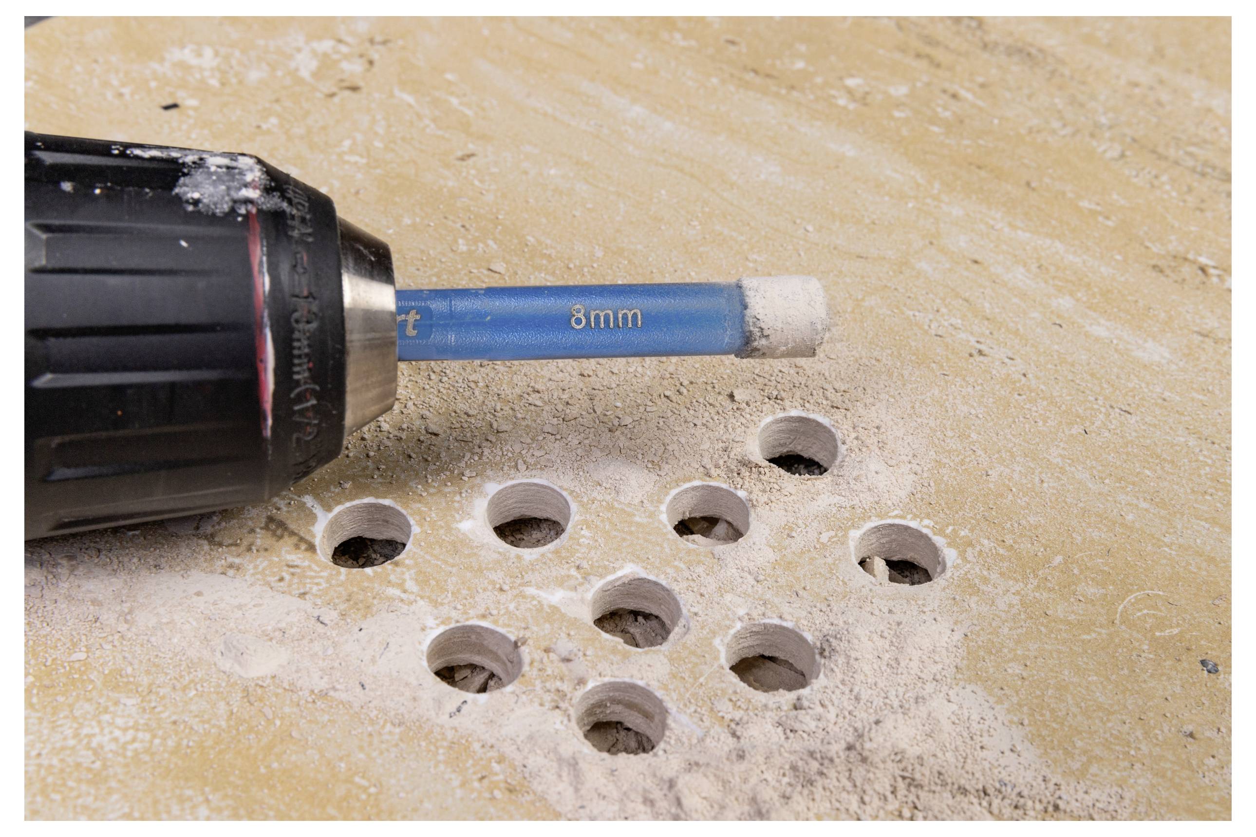 Eine Bohrmaschine mit einem 8-mm-Bohrer, der mehrere Löcher in eine beige Oberfläche bohrt, umgeben von Staub, was auf ein Holzarbeits- oder Heimwerker-Projekt hinweist.