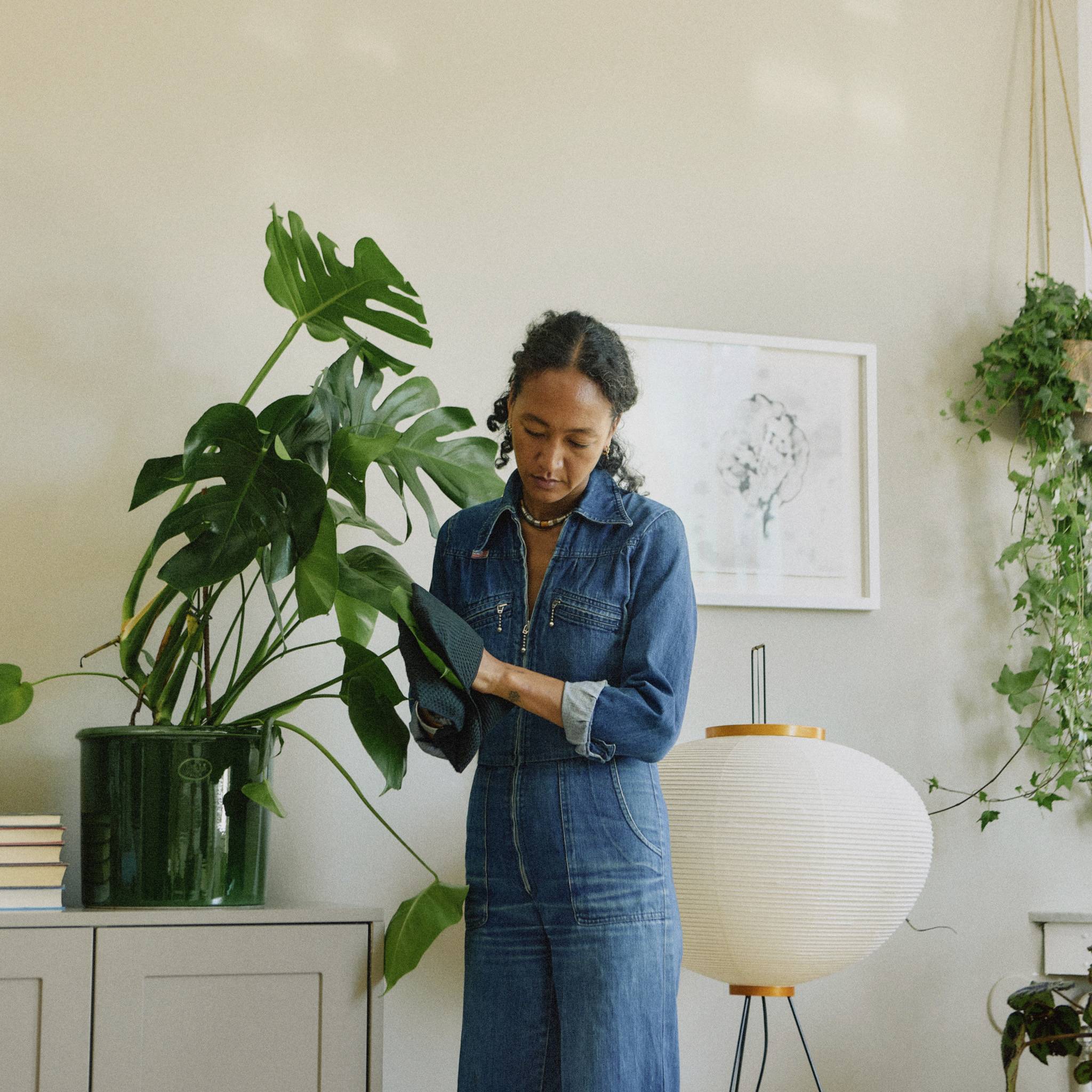 Eine Person in einem Jeans-Outfit steht neben einer großen Pflanze und einer runden Lampe, vor einem hellen Hintergrund mit Kunst an der Wand.