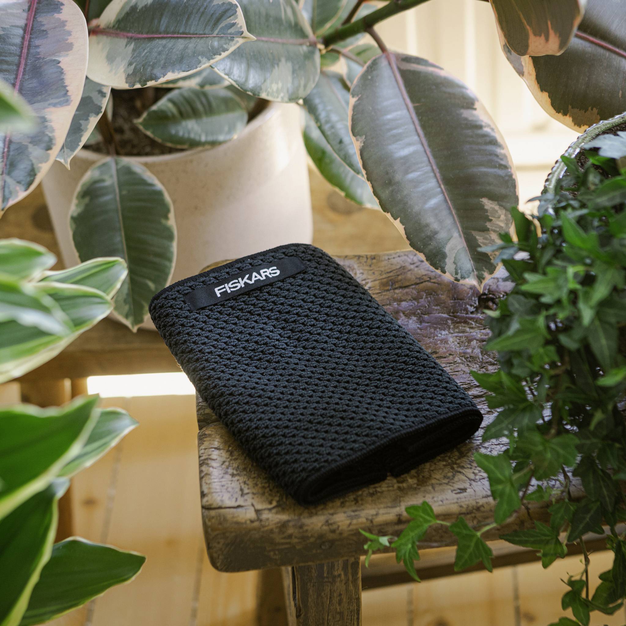 Ein schwarzes Gartenwerkzeug mit 'Fiskars'-Logo auf einem Holztisch, umgeben von verschiedenen tropischen Pflanzen in Töpfen.