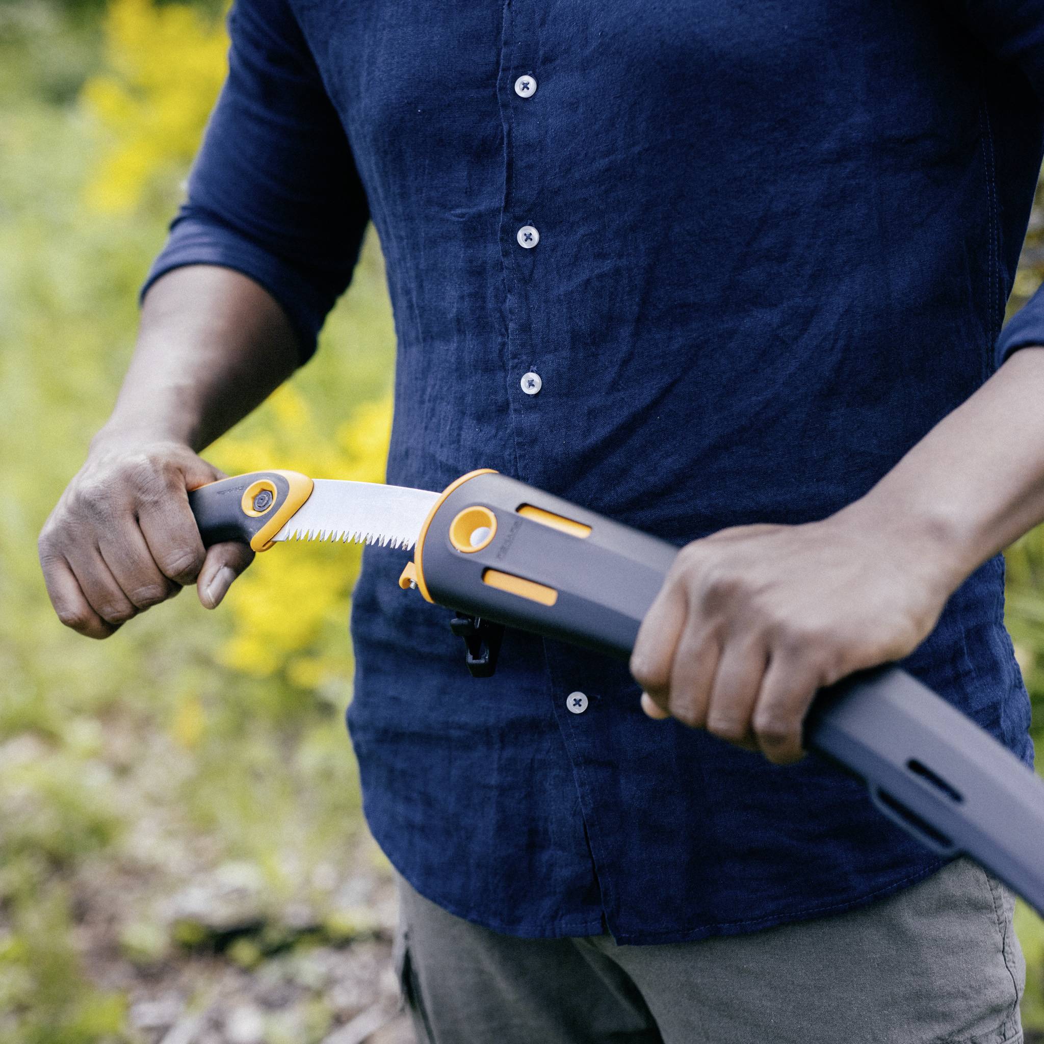 Person schneidet mit einer orange-grauen, klappbaren Handsäge im Freien einen Ast. Hintergrund zeigt unscharfe grüne Vegetation.