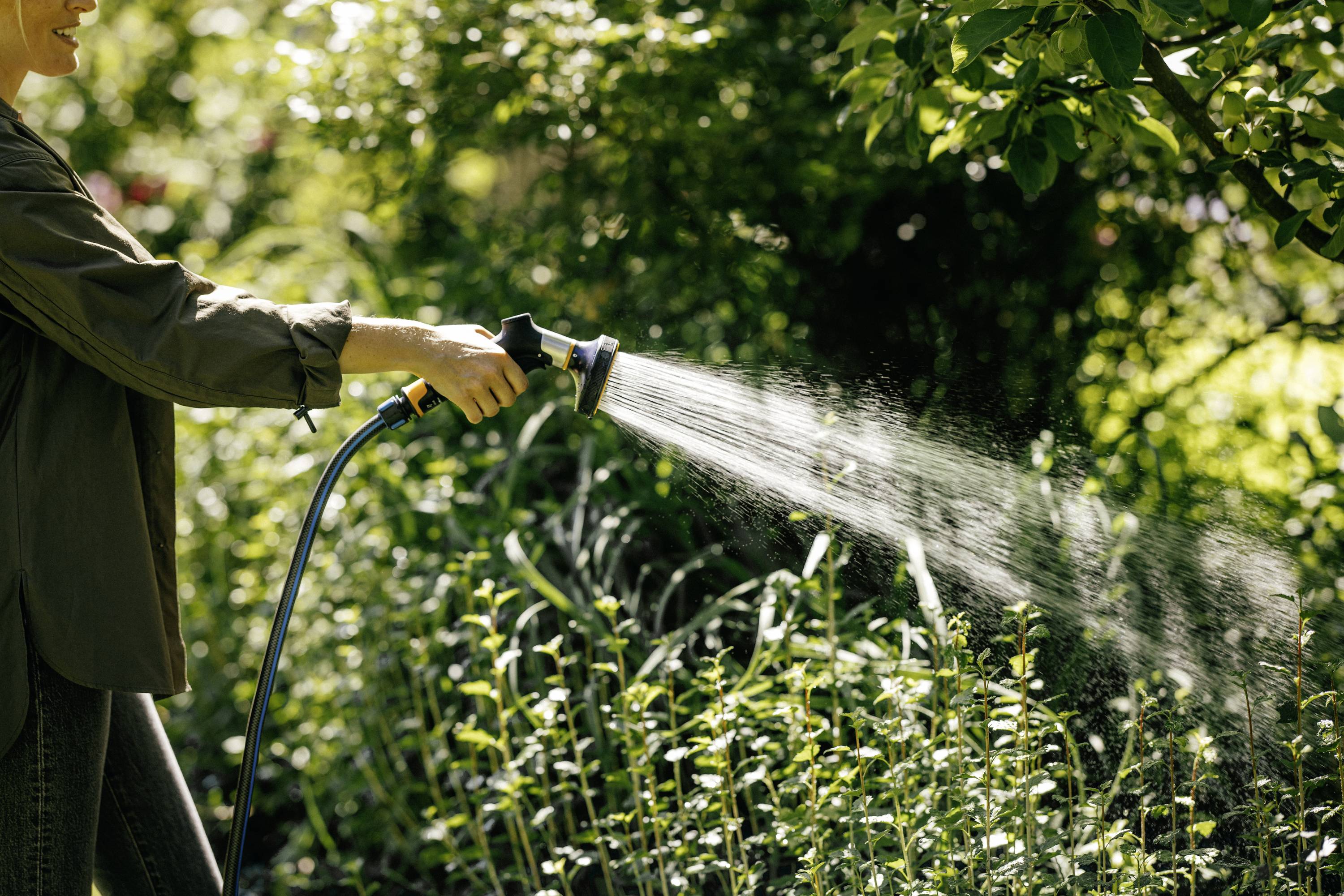 Eine Person gießt Pflanzen mit einem Gartenschlauch in einem grünen Garten. Наступає зелена трава та дерева у фоновому режимі.