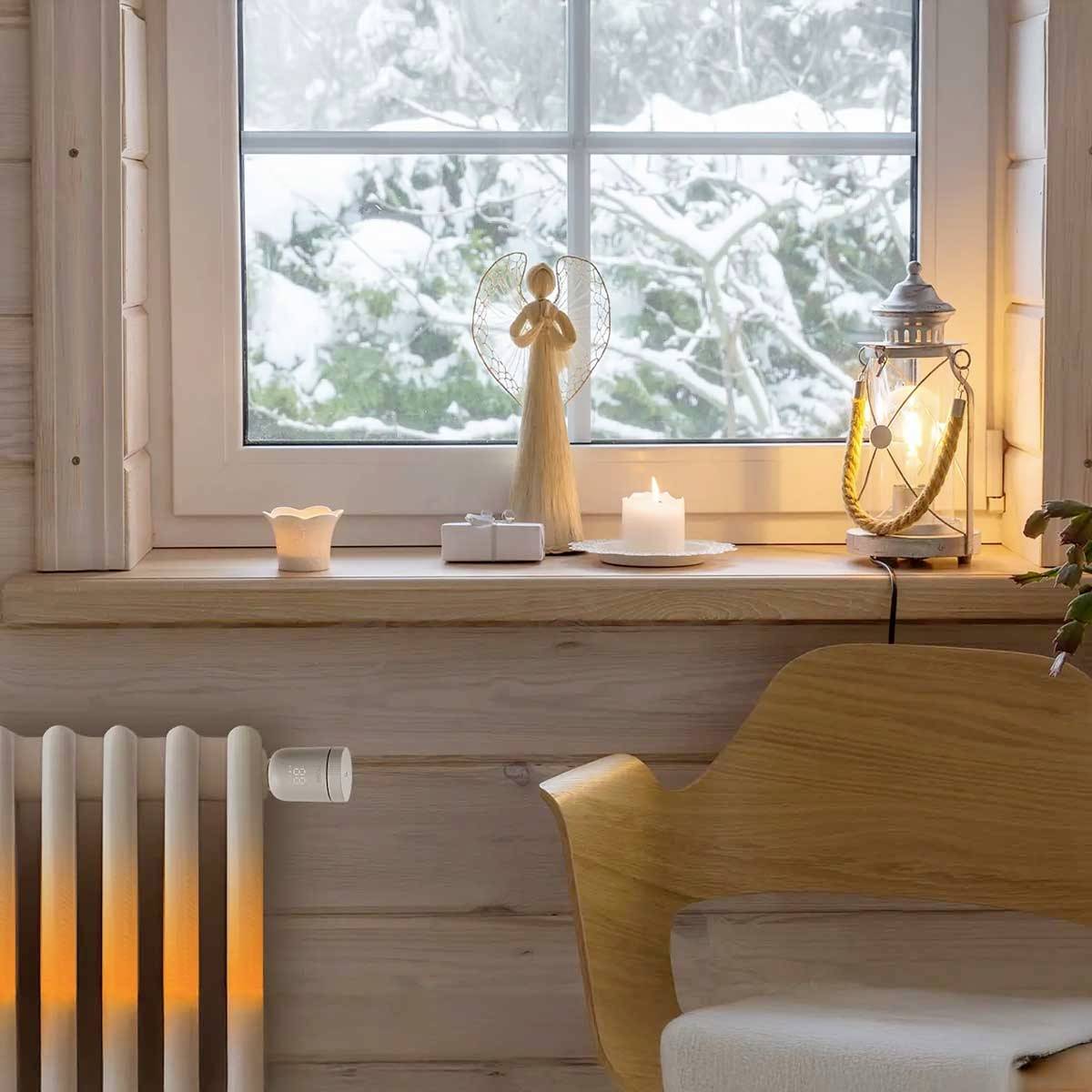 Gemütlicher Raum mit Holzofen, Lampe, Kerze und Engel auf Fensterbank; draußen schneebedeckte Landschaft sichtbar.