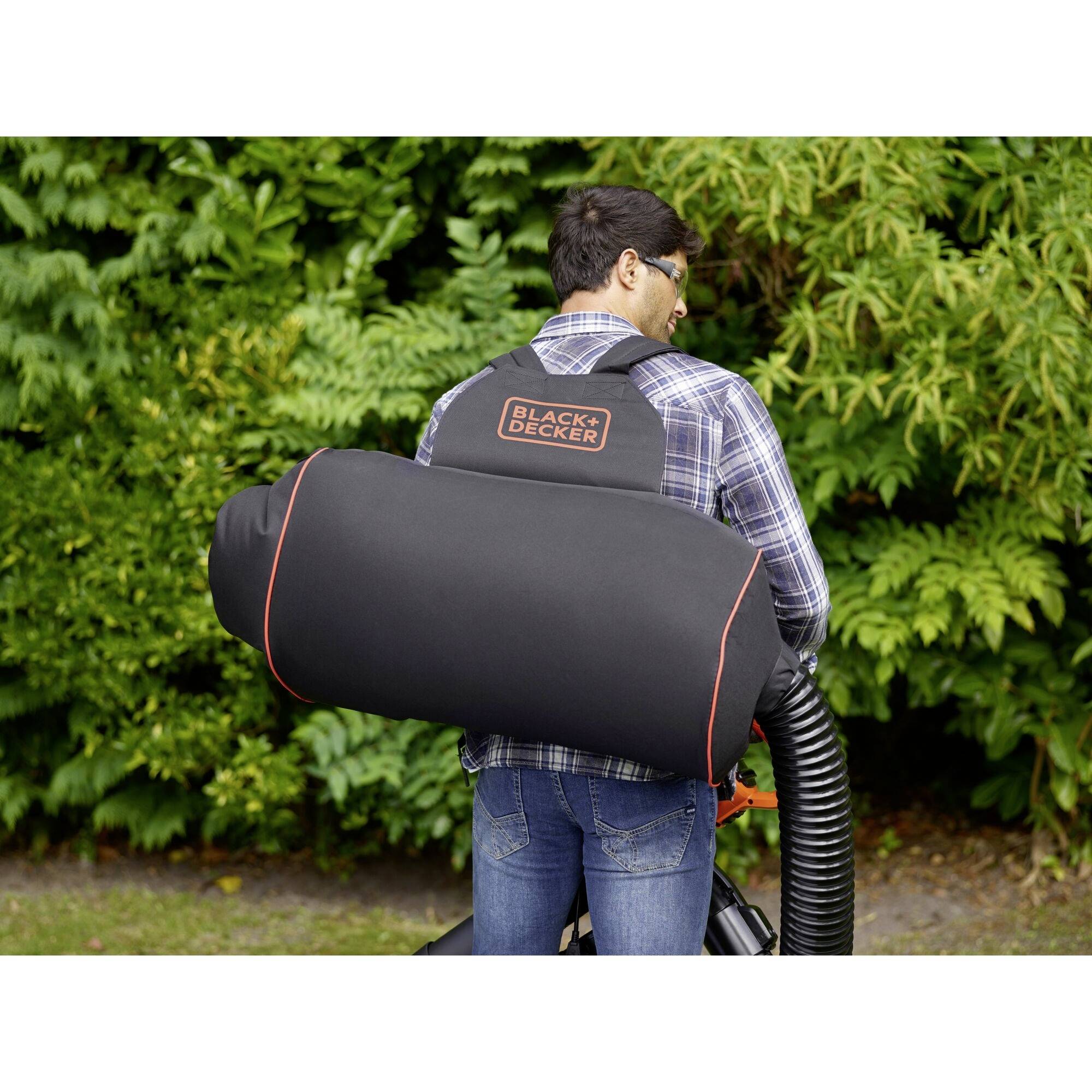 Ein Mann trägt einen schwarzen Laubsaugerrucksack von Black & Decker und saugt Laub in einem Garten mit üppigem Grün im Hintergrund ein.