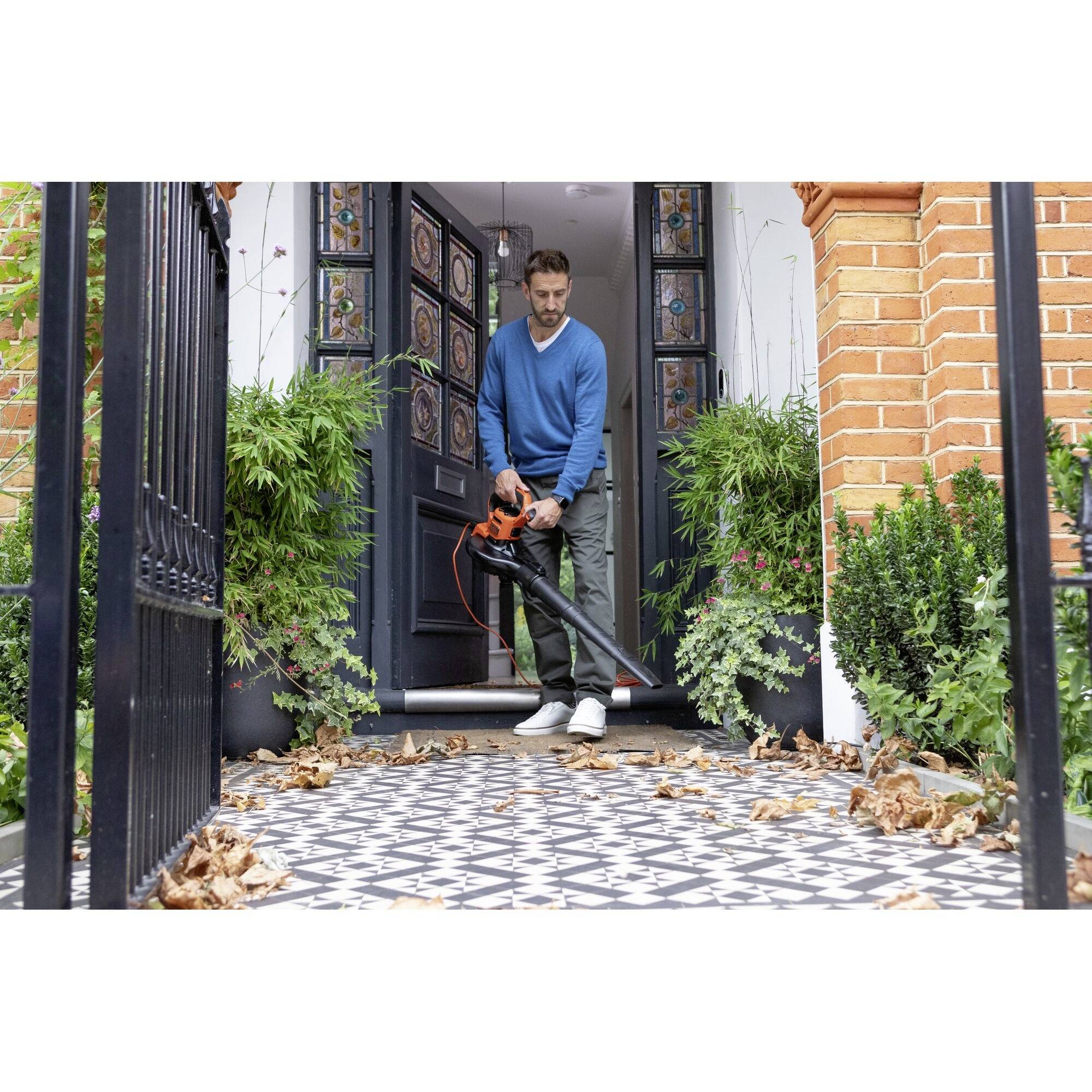 Ein Mann steht auf einer gefliesten Terrasse mit einem Laubbläser in der Hand, umgeben von Pflanzen und buntem Laub.