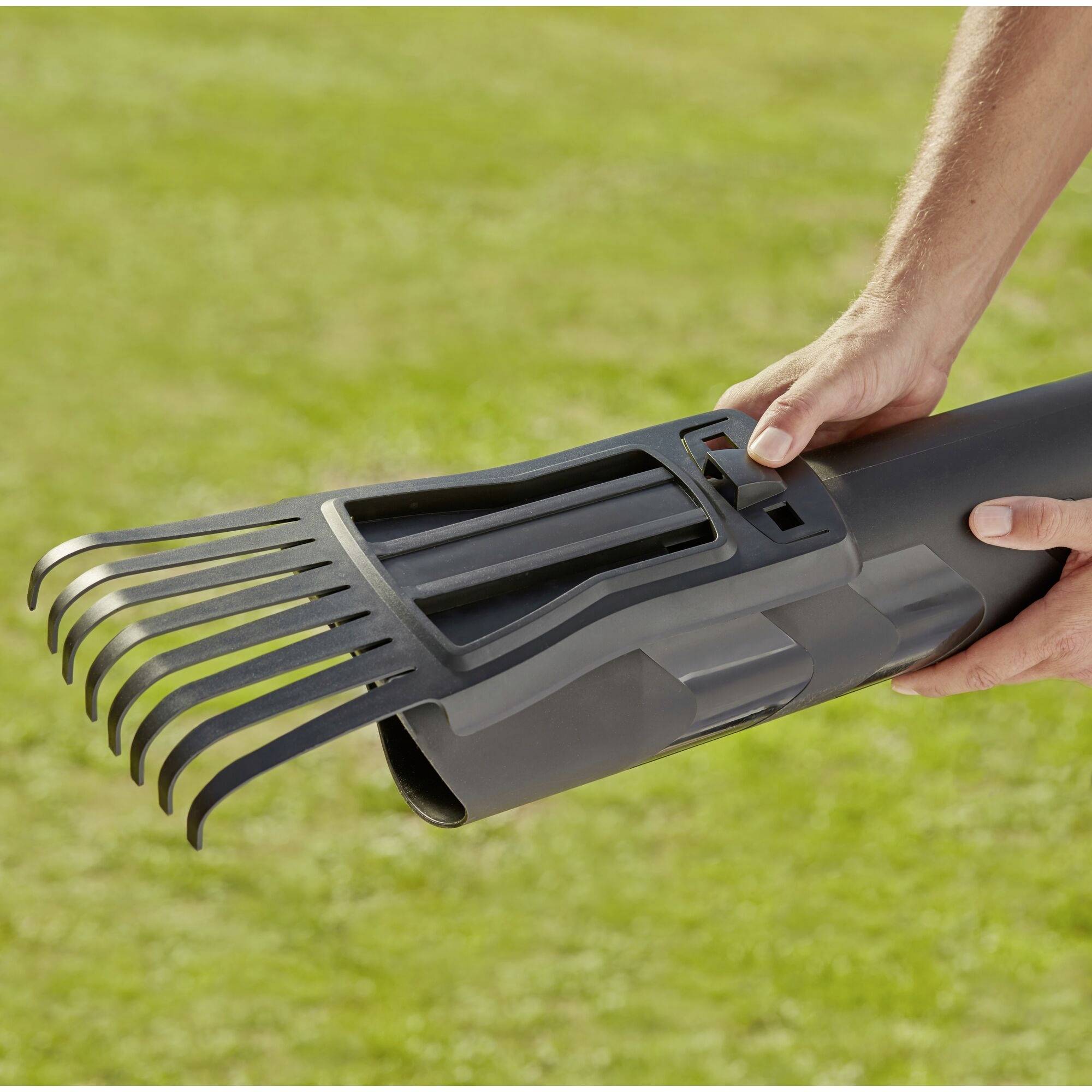 Eine Person hält ein graues Gerät mit schwarzen Kammzähnen, das wie ein Teleskop-Rohr aussieht, über einer grünen Grasfläche.