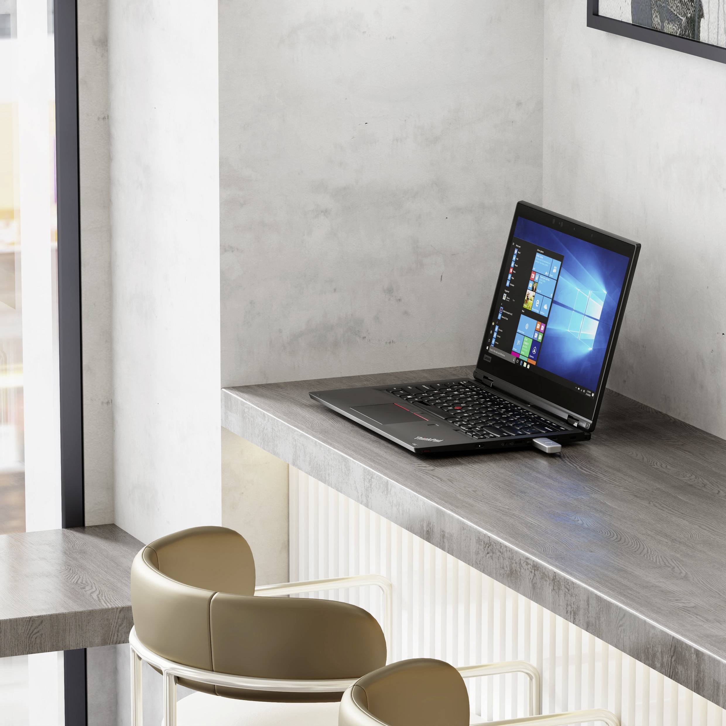 Ein Laptop mit einem Windows-Bildschirm steht auf einem kleinen grauen Schreibtisch in der Nähe eines Fensters, neben einem hellgefärbten Stuhl in einem modernen, minimalistischen Raum.