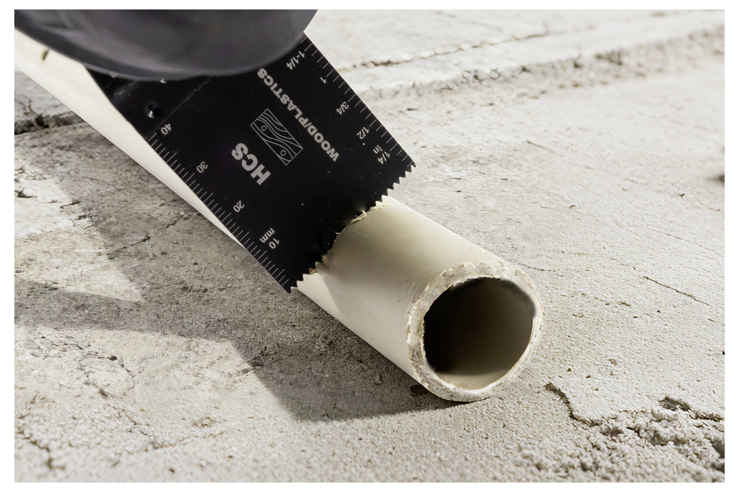 Eine Handsäge schneidet durch ein weißes PVC-Rohr, das auf einer rauen Betonoberfläche liegt.