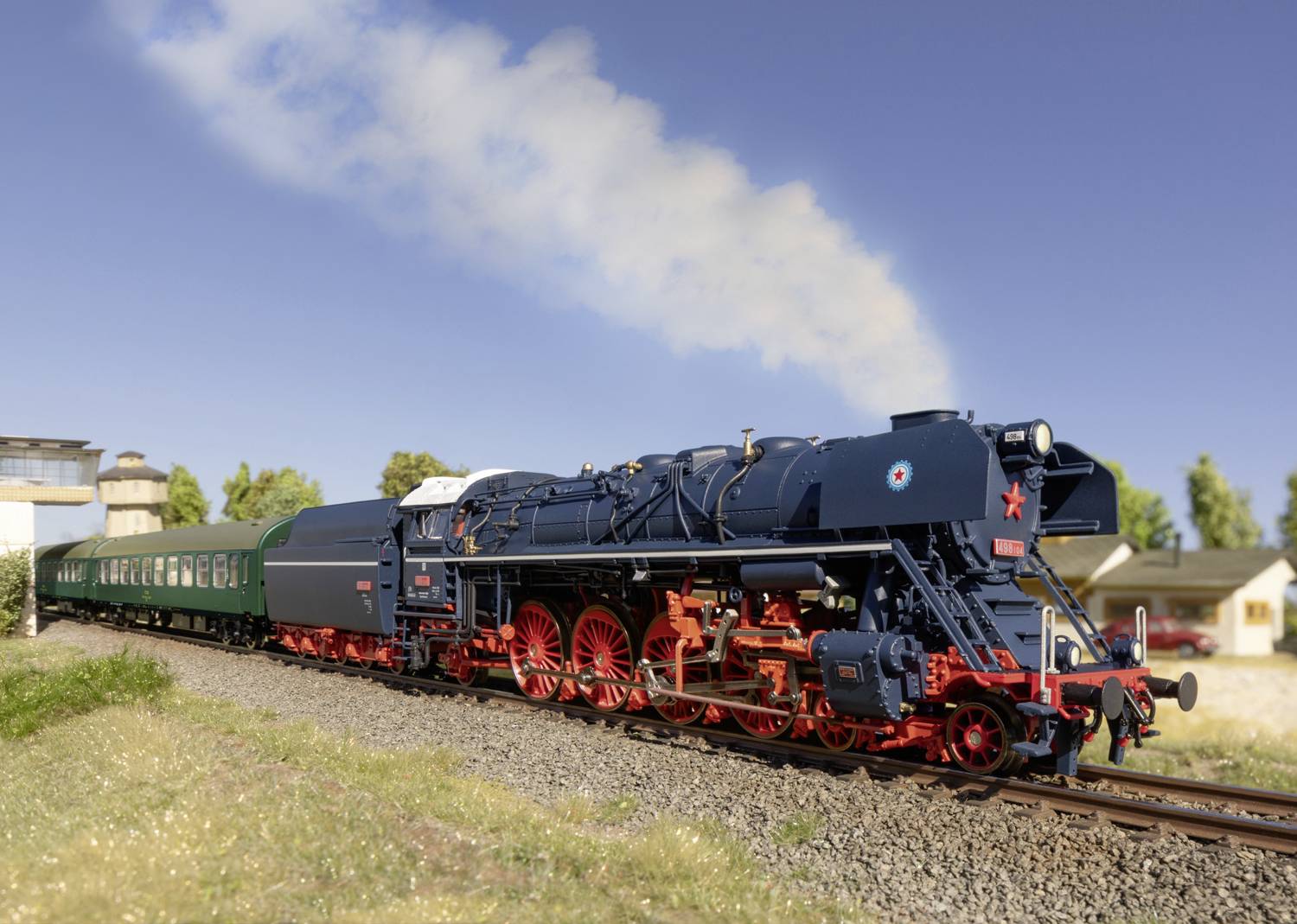 Eine Modellbahn-Dampflokomotive zieht mehrere grüne Waggons auf einem Gleis durch eine ländliche Landschaft bei klarem Himmel.
