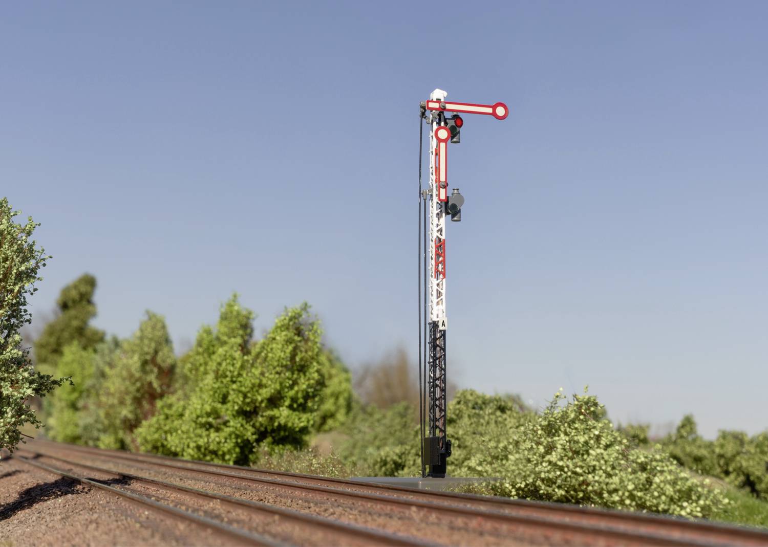 Ein mechanisches Eisenbahnsignal zeigt 'Halt' an einer Bahngleise, umgeben von Büschen unter einem klaren blauen Himmel.