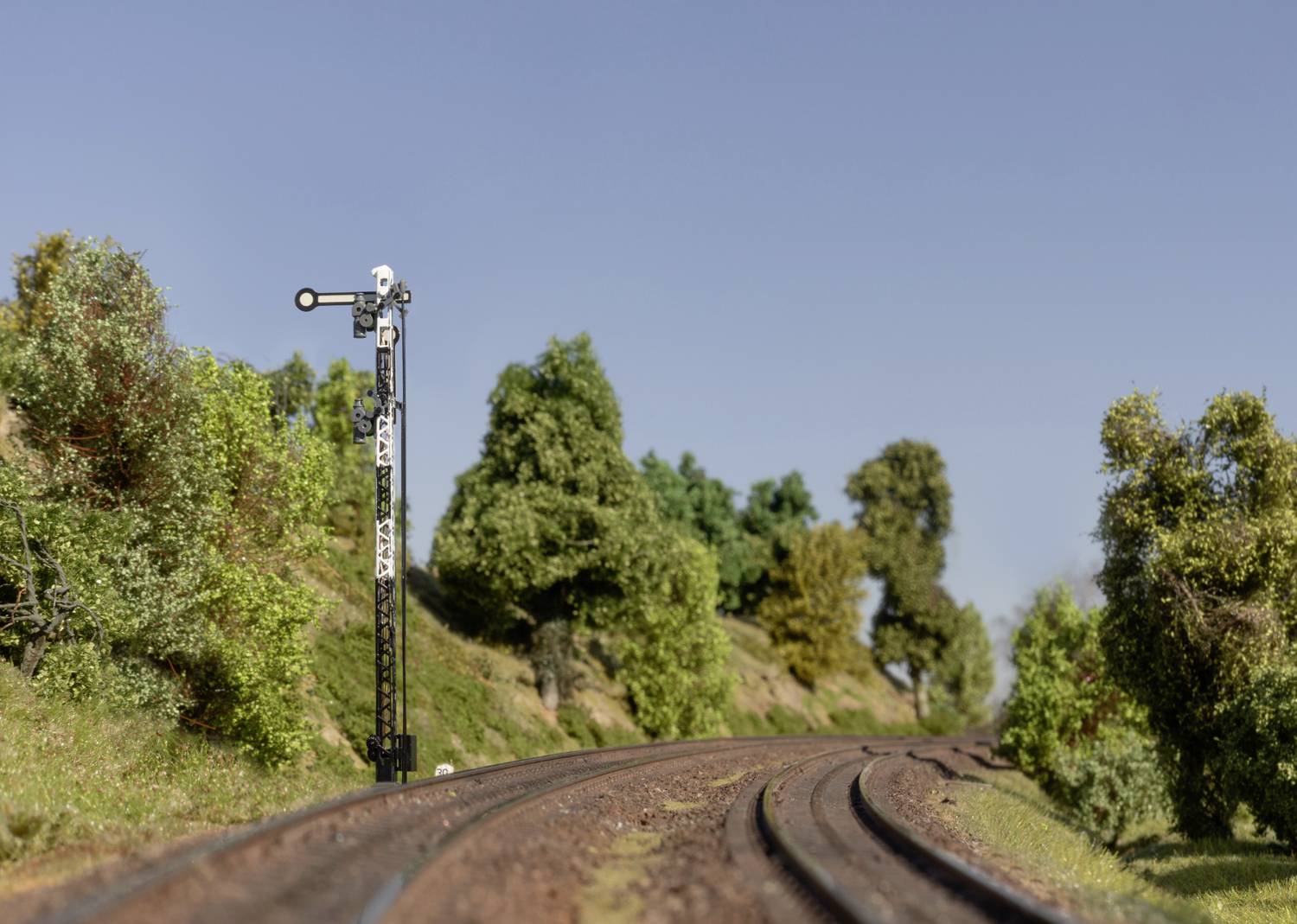 Eisenbahnsignal neben zweigleisiger Eisenbahnstrecke, umgeben von Hügeln und Bäumen, unter klarem Himmel.