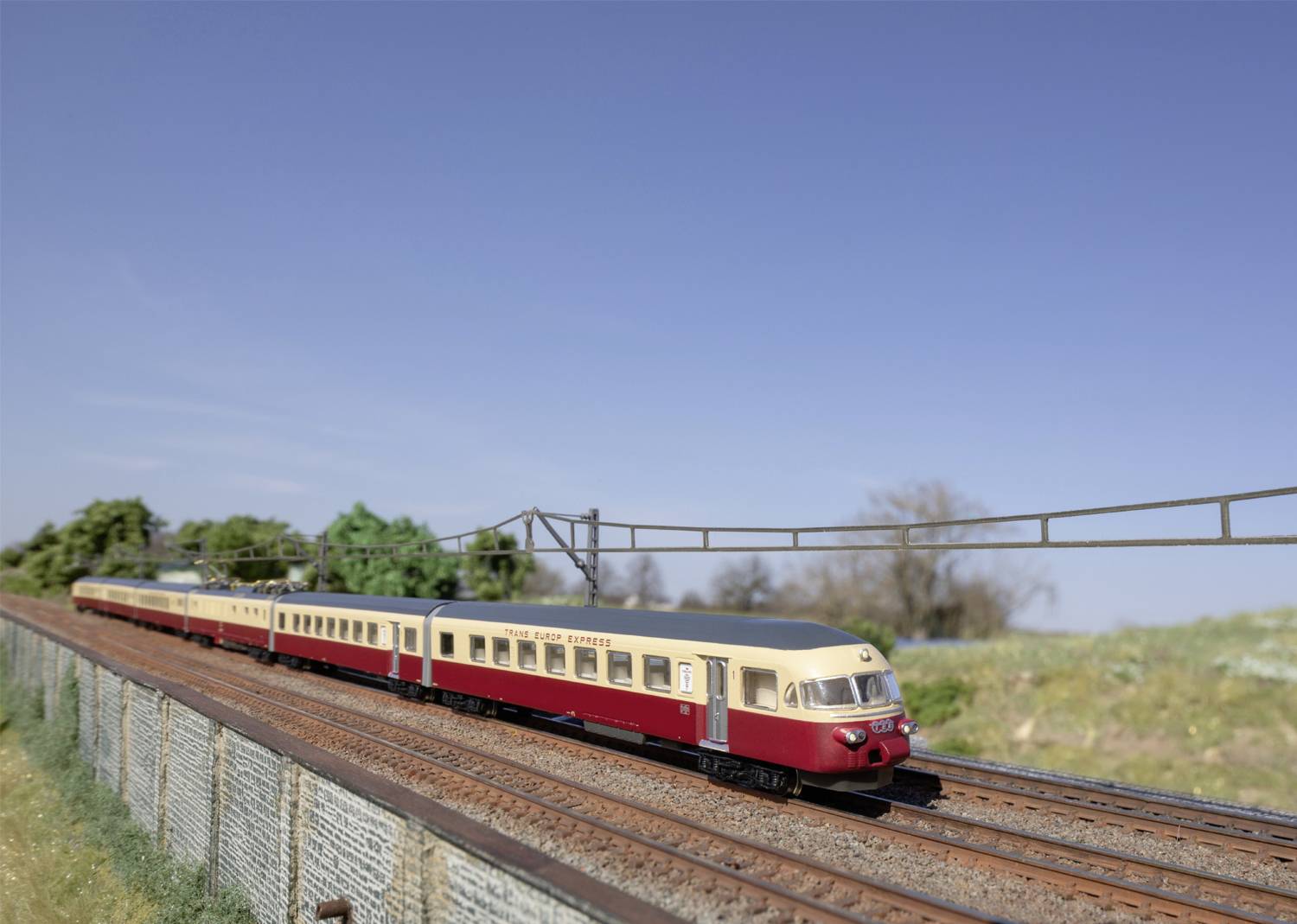 Zugmodell auf einer Miniatur-Eisenbahnstrecke mit Strommasten, umgeben von Landschaft mit Bäumen und klarem Himmel im Hintergrund.