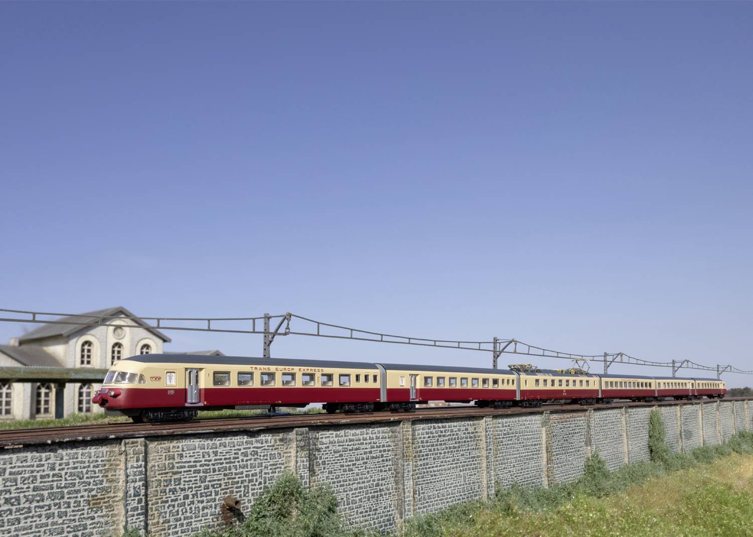 Ein roter Zug fährt über eine erhobene Bahnstrecke neben einem Gebäude und unter einem klaren blauen Himmel.