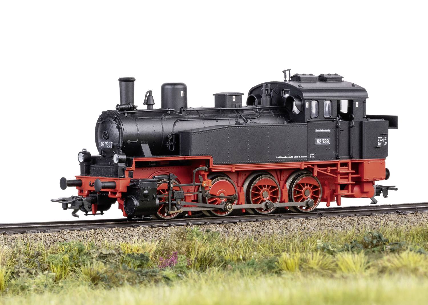 Eine schwarze Dampflok mit rotem Unterbau steht auf Bahngleisen in einer Wiesenlandschaft, Modell 130.7716.