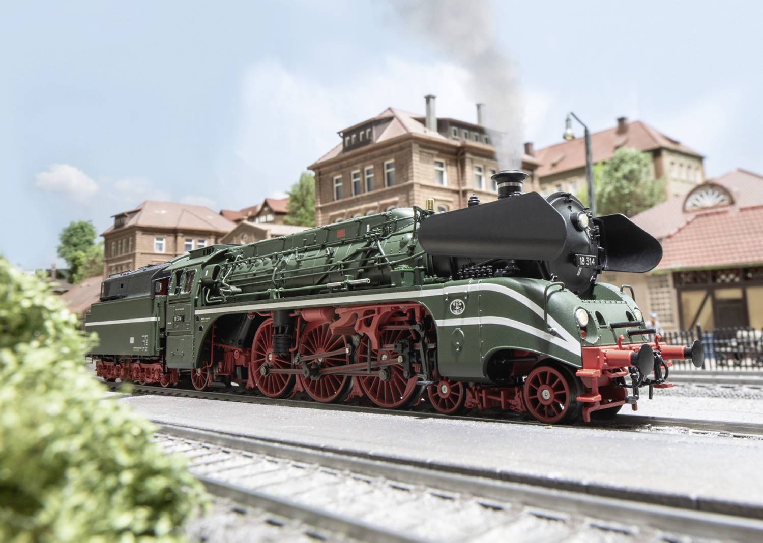 Grüner und roter Dampfzug fährt auf Gleis vorbei an historischen Gebäuden unter blauem Himmel. Rauch steigt aus dem Schornstein.