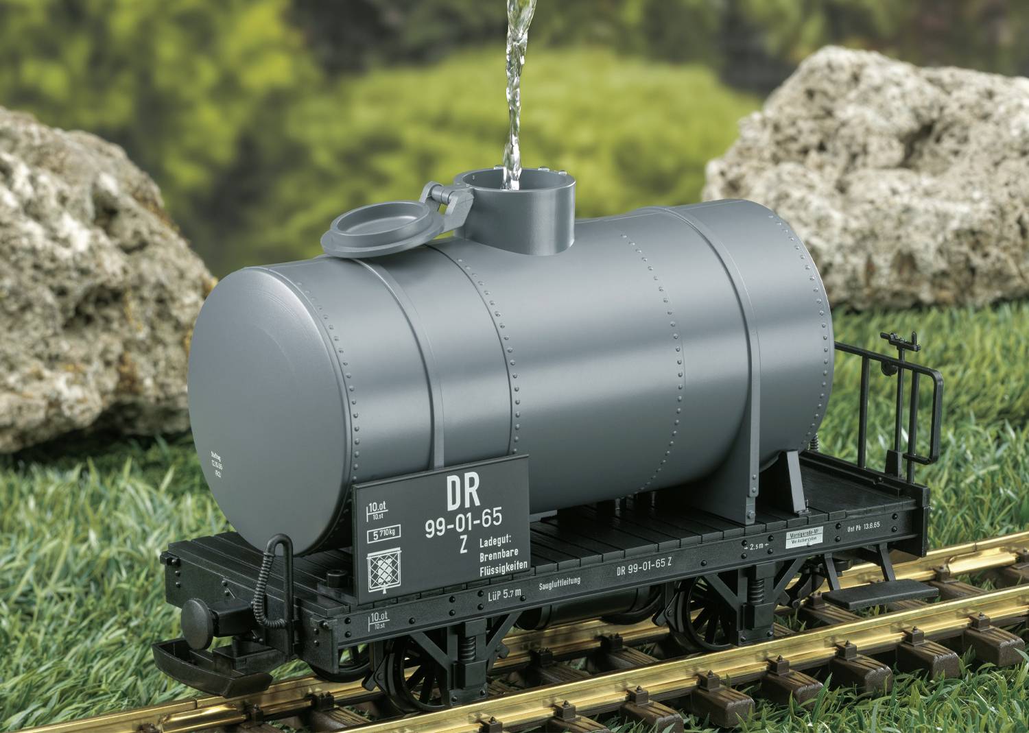Grauer Modell-Eisenbahnkesselwagen mit geöffneter Klappe, durch die Wasser eingefüllt wird; umgeben von Gras und Felsen im Hintergrund.