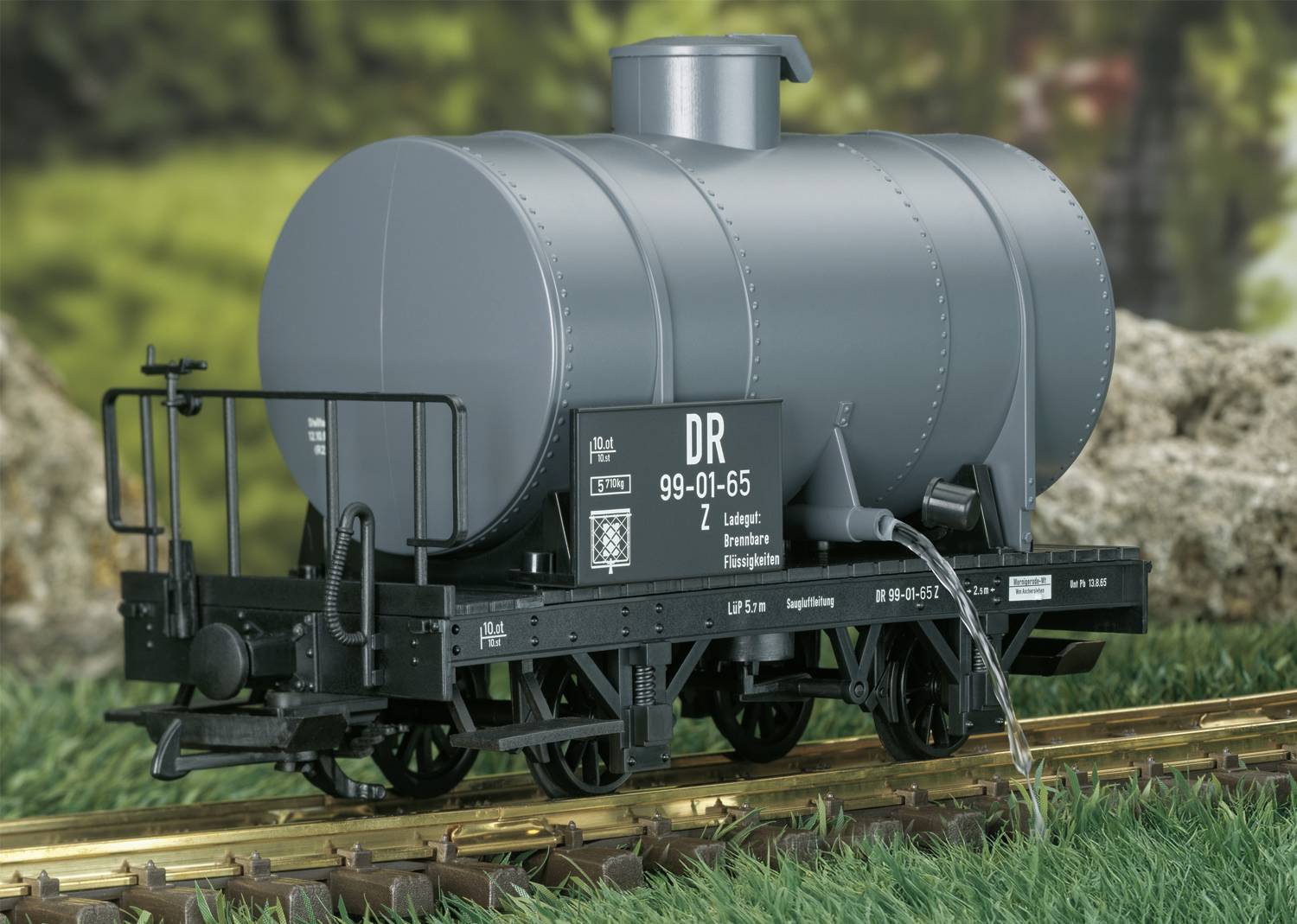 Ein grauer Modell-Eisenbahnkesselwagen auf einem Gleis, dargestellt in einer natürlichen Umgebung mit Gras und Felsen im Hintergrund.