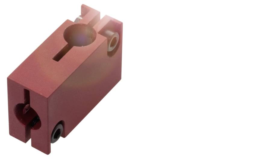 Ein roter Metallblock mit mehreren Löchern und Schlitzen, vermutlich ein Maschinenteil oder Befestigungselement.
