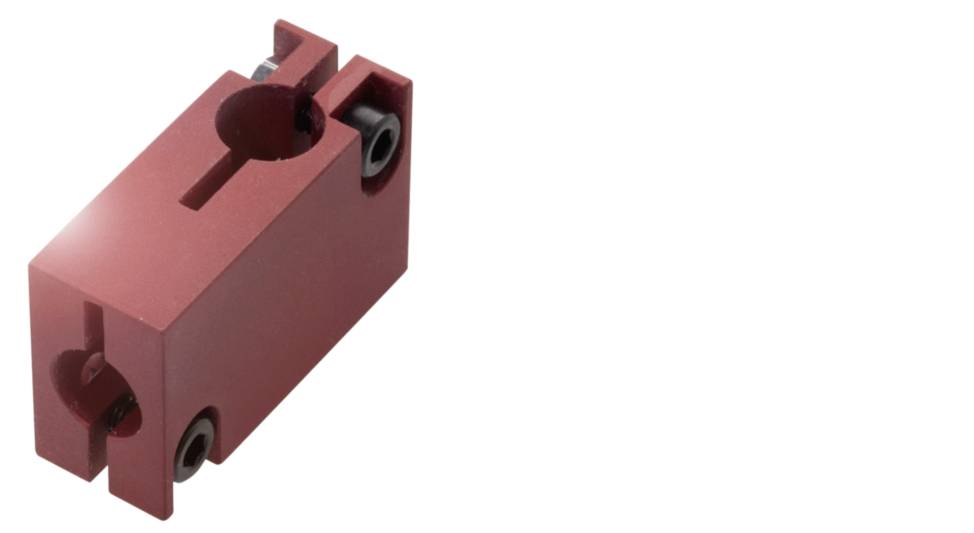 Ein roter, rechteckiger Metallblock mit zwei kreisförmigen Vertiefungen und Schraubenlöchern an den Seiten, möglicherweise ein mechanisches Bauteil.