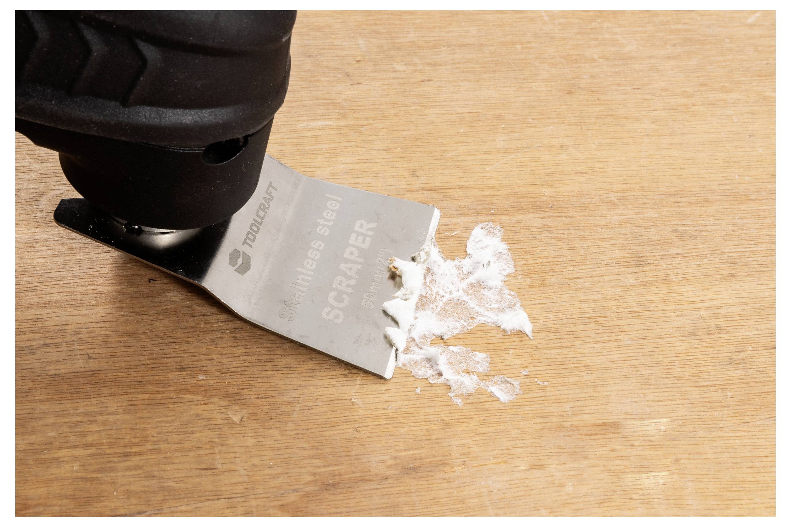 Ein Metallschaber entfernt weiße Farbresiduen von einer Holzoberfläche und unterstreicht dabei die Wirksamkeit des Werkzeugs beim Reinigen und Vorbereiten von Oberflächen.