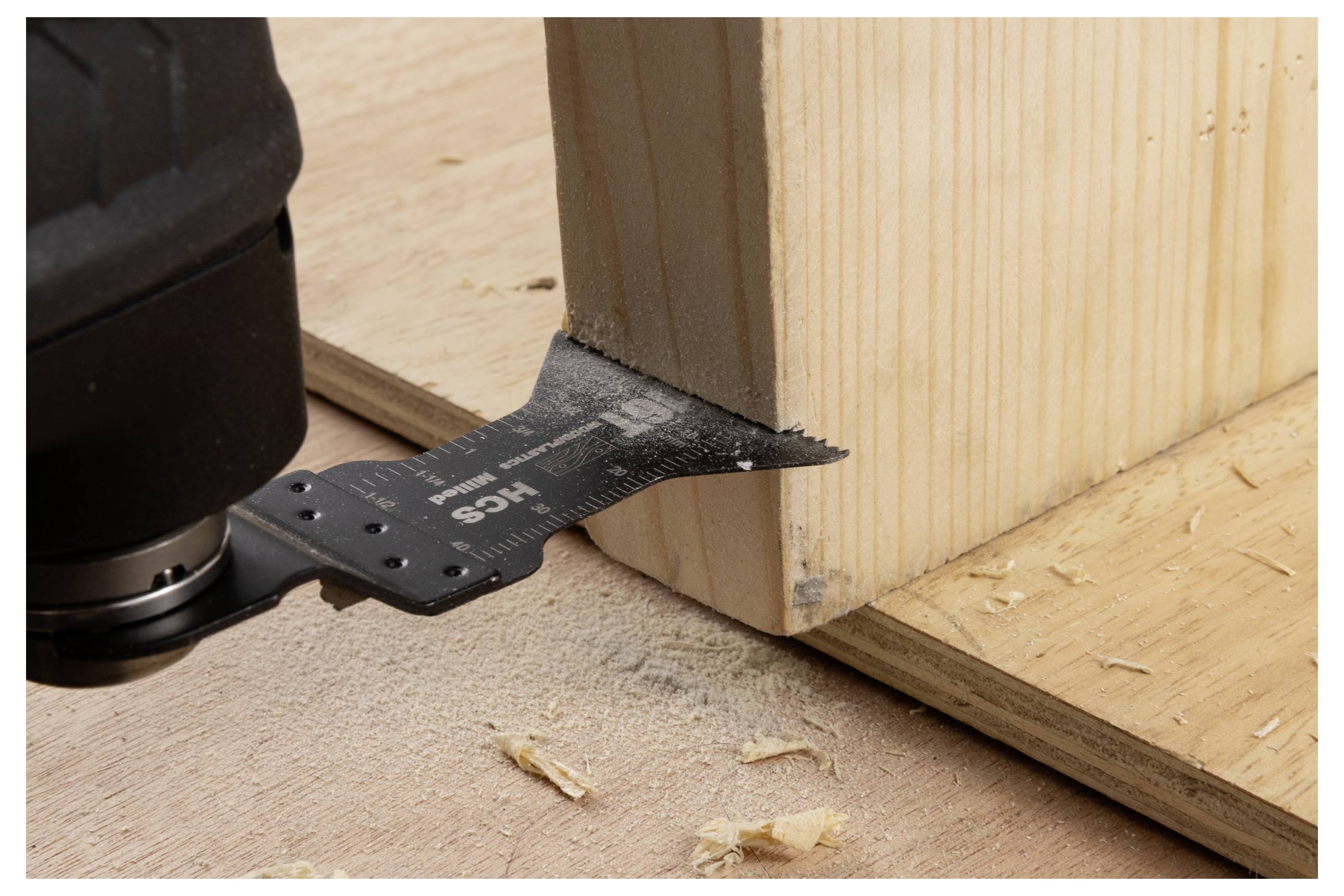 Eine Nahaufnahme eines Multitools mit einem Sägeblatt-Aufsatz, der durch eine Holzplatte schneidet, mit Holzspänen auf der Oberfläche drumherum.