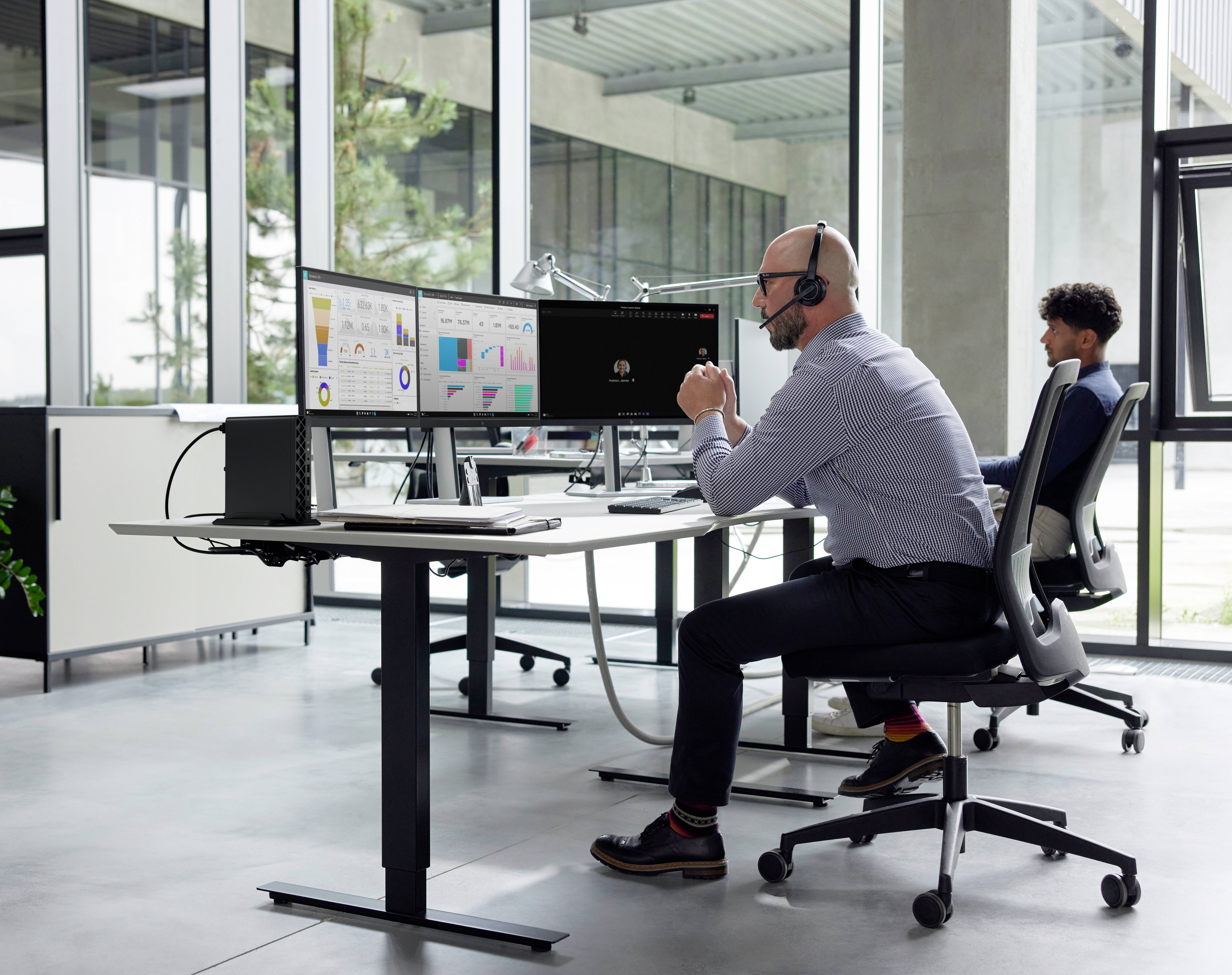 Ein Mann mit Headset arbeitet an einem Schreibtisch mit mehreren Monitoren, auf denen Graphen zu sehen sind, während eine andere Person an einem nahegelegenen Schreibtisch im Büro sitzt.