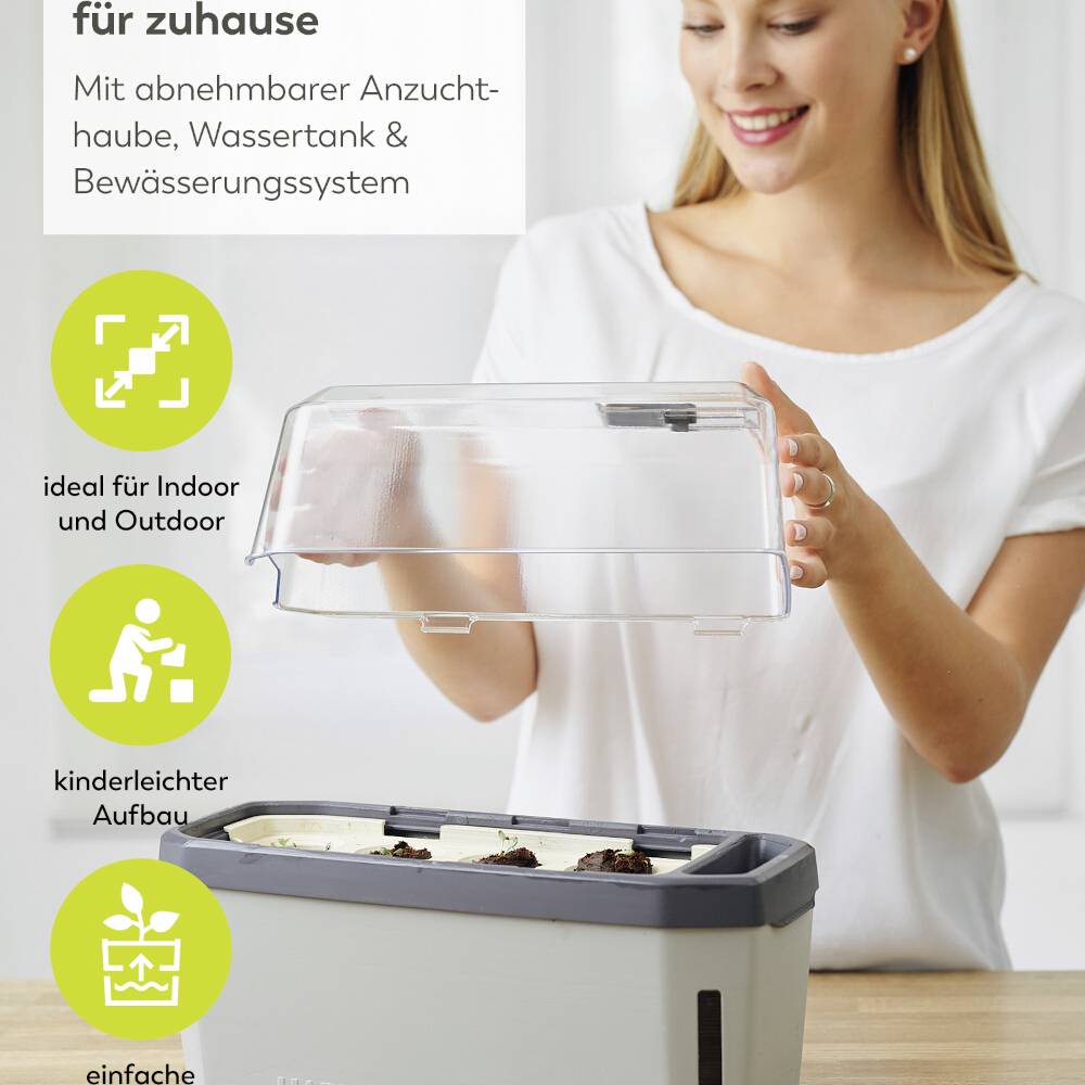 Frau zeigt Mini-Gewächshaus für drinnen und draußen mit abnehmbarer Haube, Wassertank und Bewässerungssystem.