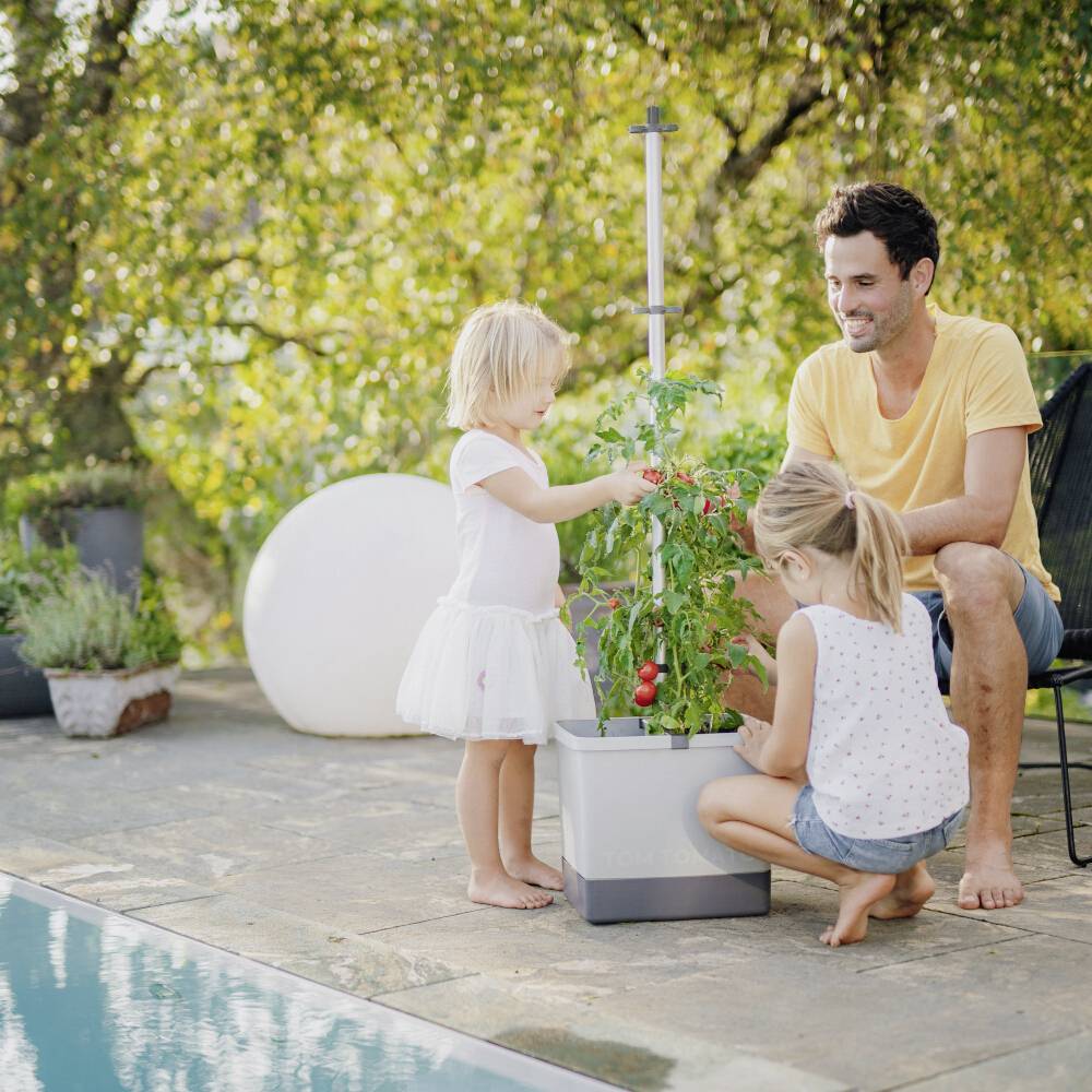 Ein Mann und zwei Kinder gießen Pflanzen neben einem Pool. Die Szene vermittelt eine friedliche, sommerliche Atmosphäre im Freien.