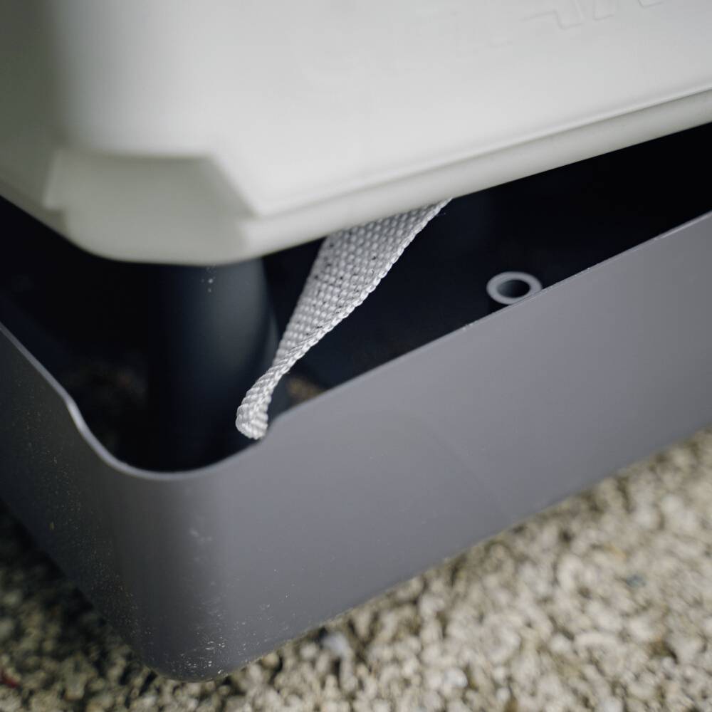 Ein grauer Behälter mit Gießkanne entleert Flüssigkeit in eine darunter liegende Auffangwanne, auf einer mit Kies bedeckten Oberfläche.