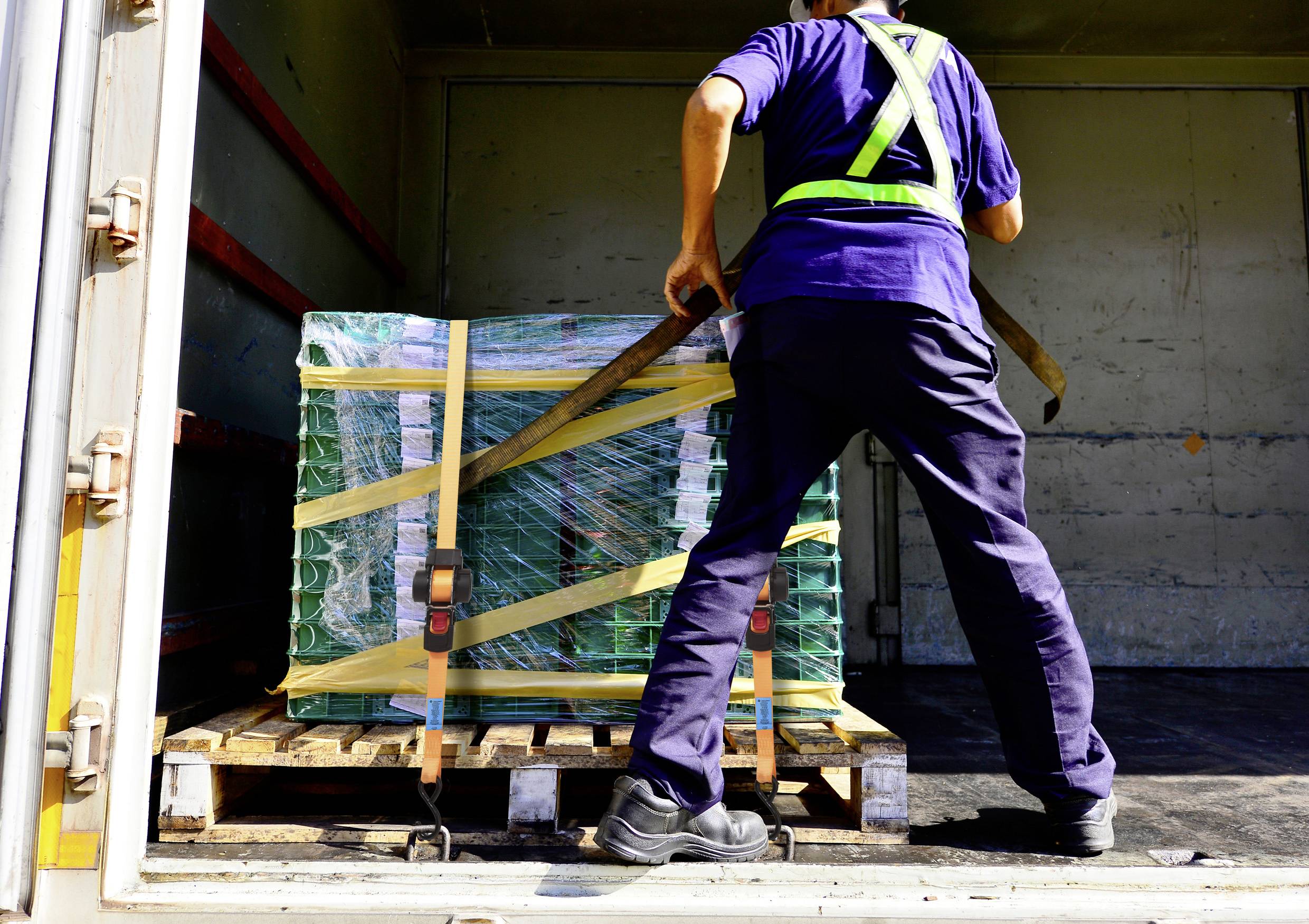 Ein Arbeiter lädt eine große, grün umwickelte Palette mit einem Hubwagen auf einen Lastwagen und trägt dabei eine Warnweste und Arbeitskleidung.