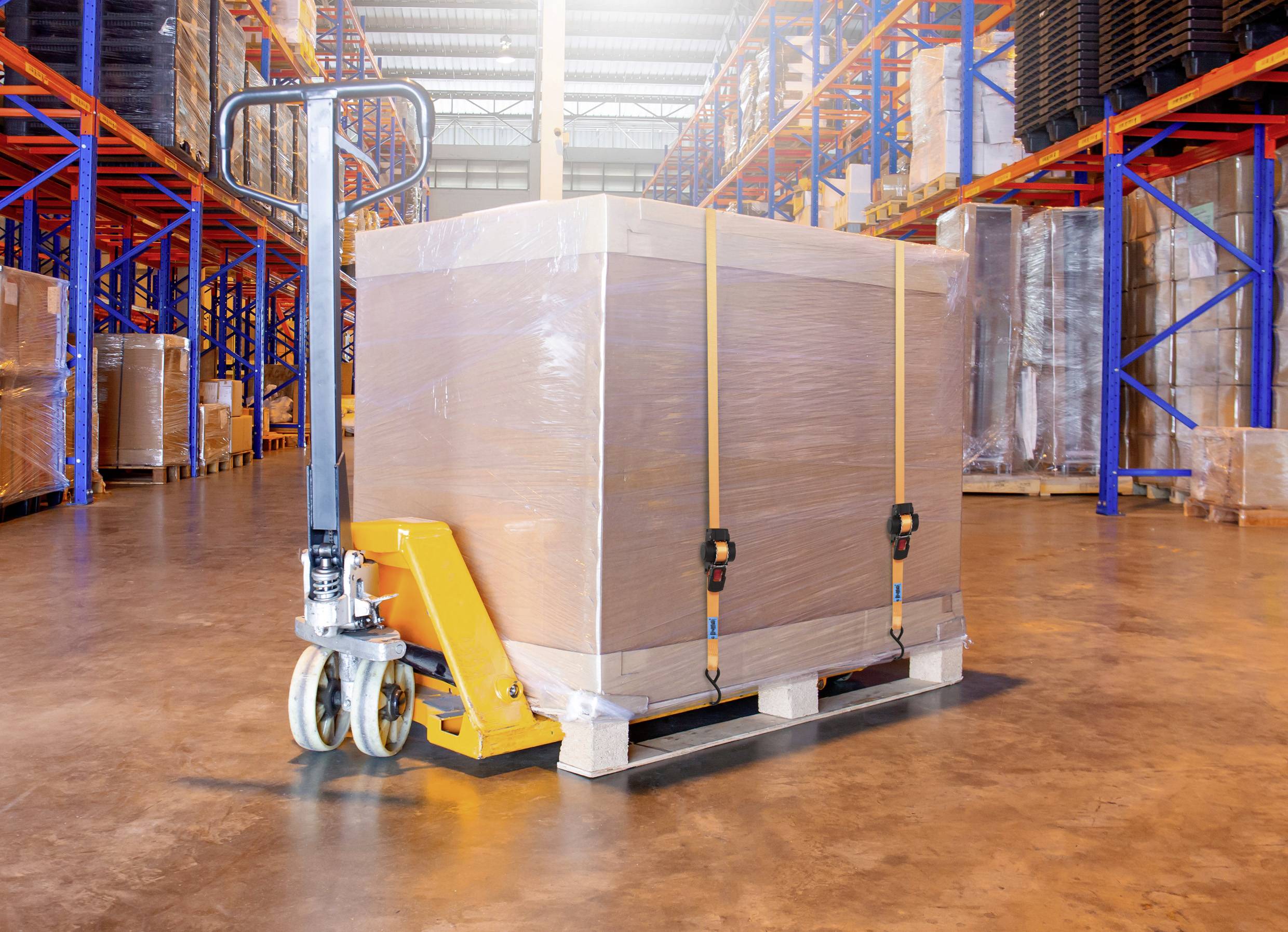 Ein Lagerraum mit einem Hubwagen, der ein großes, in Folie eingewickeltes Paket auf einer Holzpalette transportiert. Im Hintergrund sind Regale mit gestapelten Waren zu sehen.