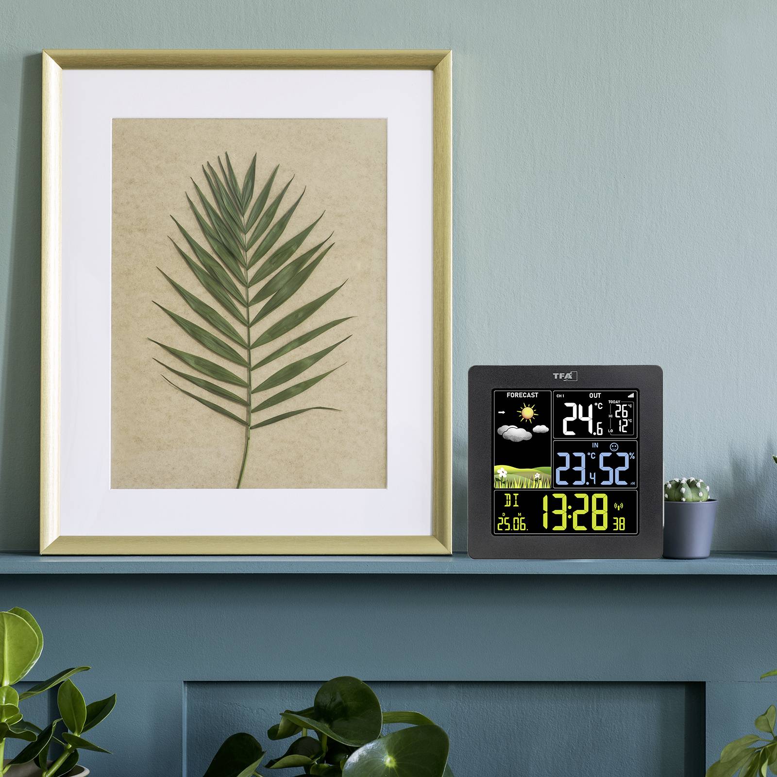 Ein digitaler Wetterstation zeigt Temperatur, Luftfeuchtigkeit und Uhrzeit an. Daneben steht ein Bild mit einem grünen Palmenblatt.