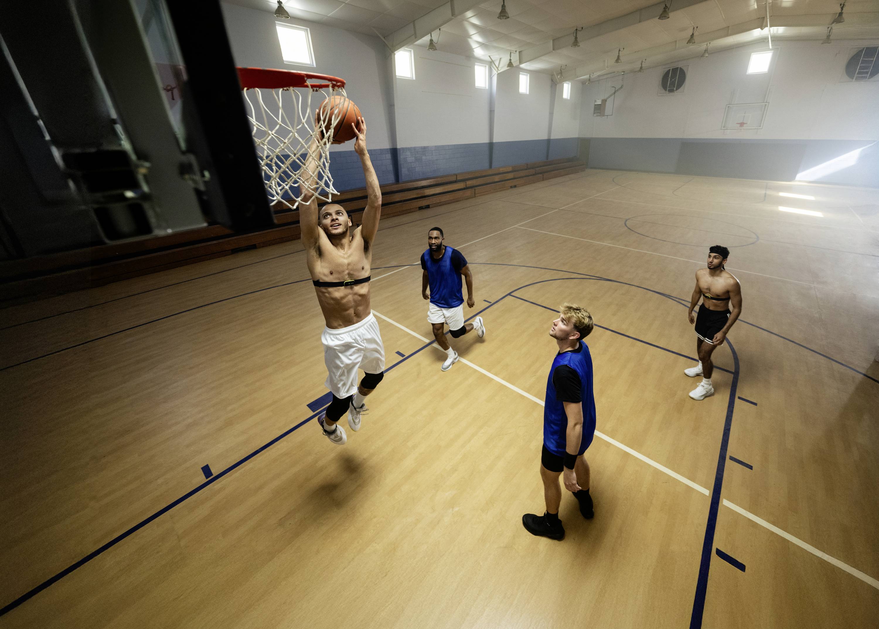 Ein Basketballspieler springt zum Dunking, während drei andere Spieler zuschauen. Sie befinden sich in einer leeren Sporthalle.