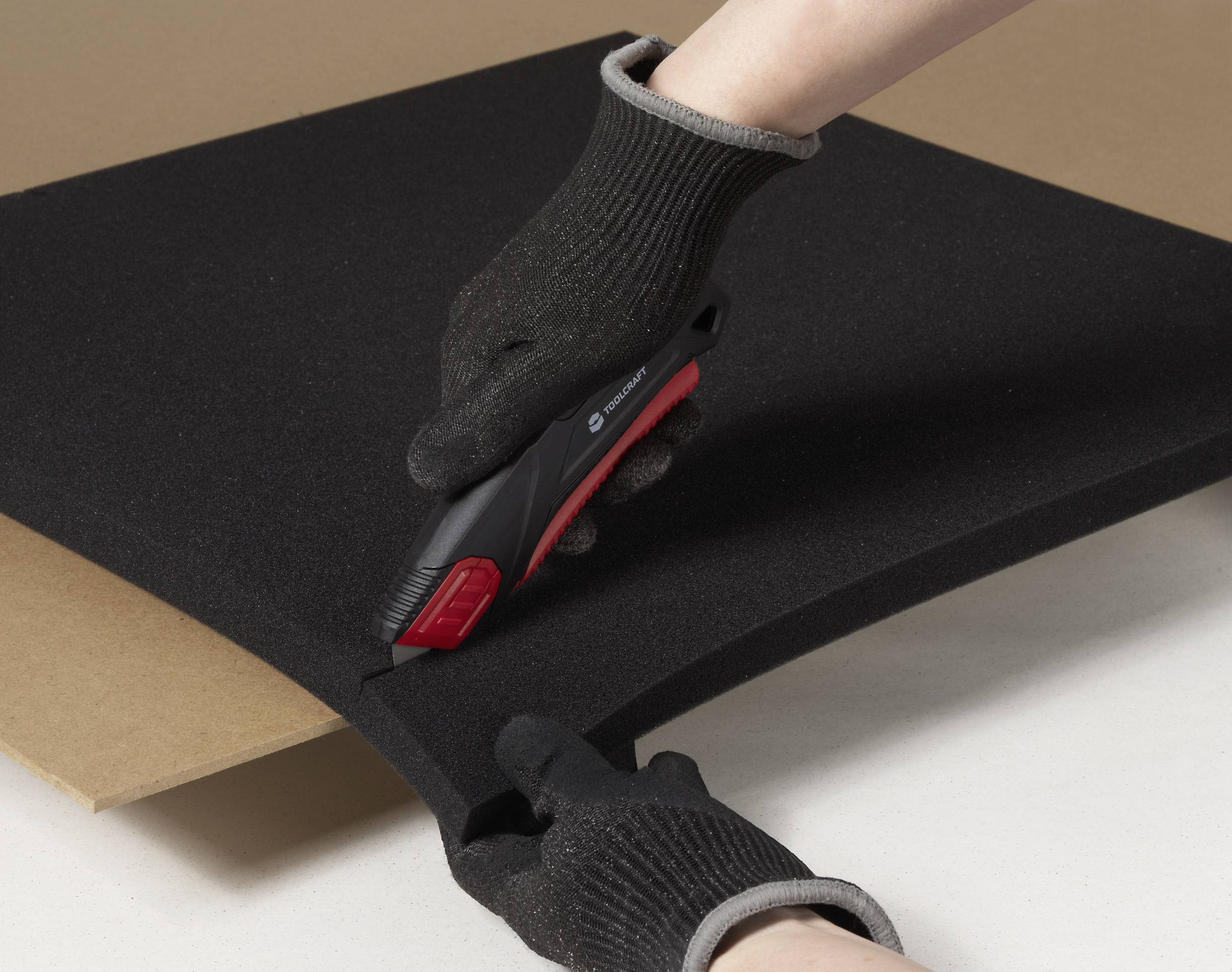 Eine Person mit Handschuhen schneidet ein dickes schwarzes Schaumblatt mit einem Universalmesser auf einer flachen Oberfläche.