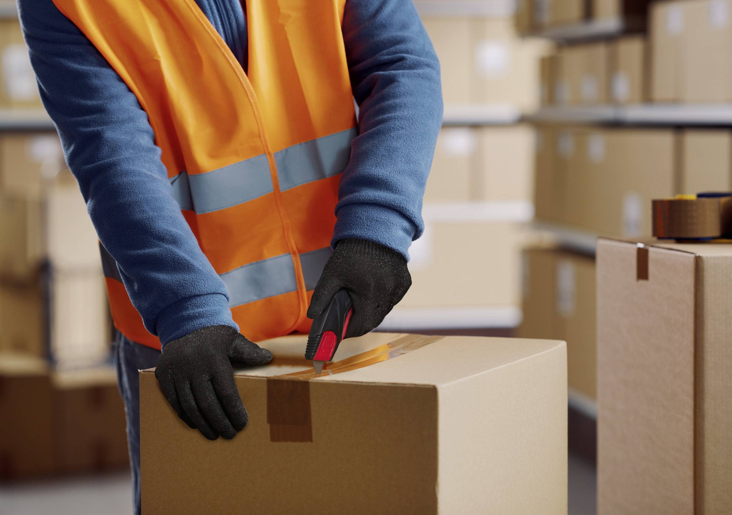Eine Person mit einer orangefarbenen Sicherheitsweste und Handschuhen verwendet einen Teppichmesser, um in einem Lagerhaus mit Regalen voller Kartons eine Pappschachtel zu öffnen.