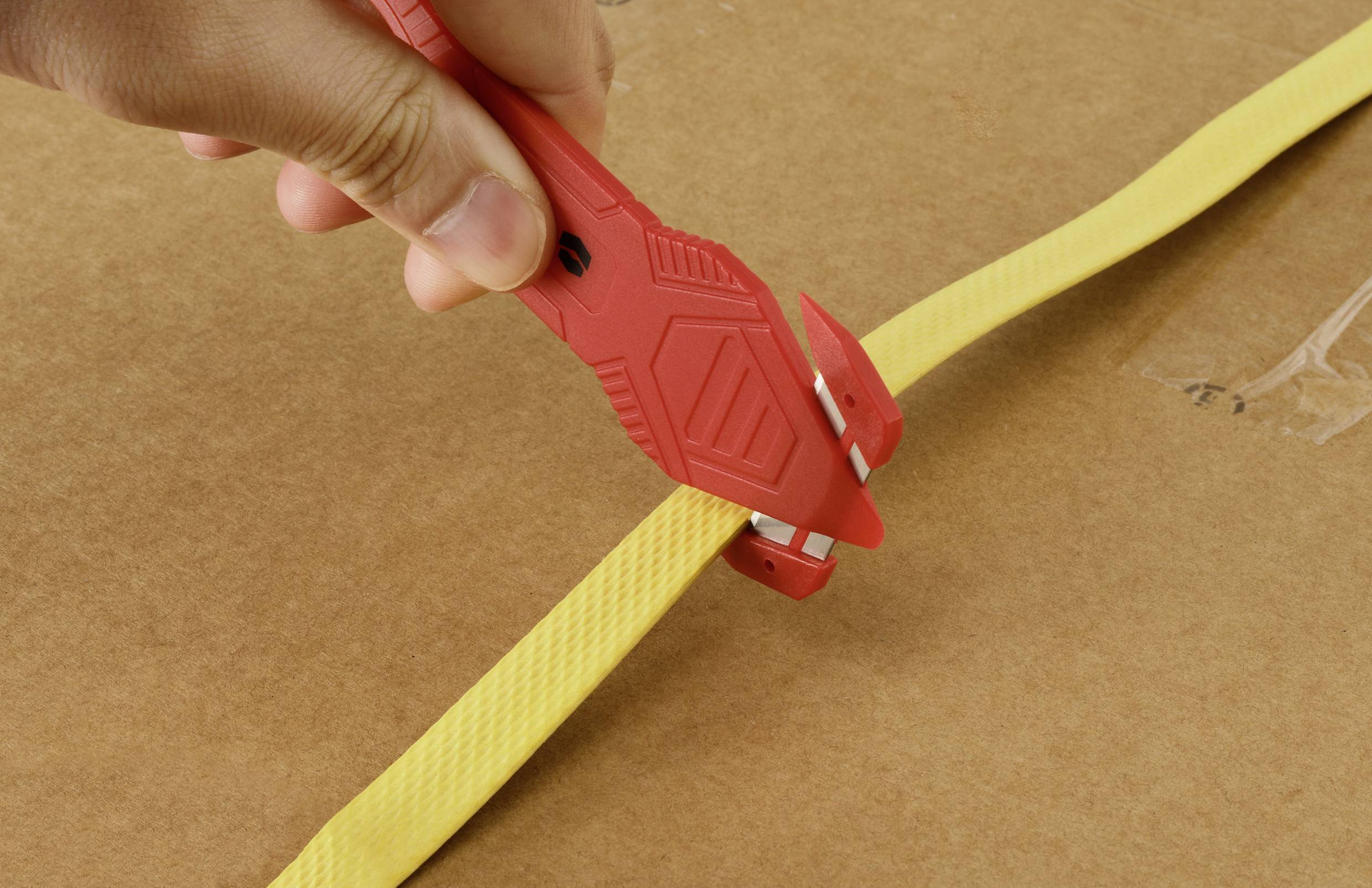 Eine Hand, die einen roten Sicherheitsmesser verwendet, um einen gelben Gurt auf einer Kartonschachtel zu durchtrennen.