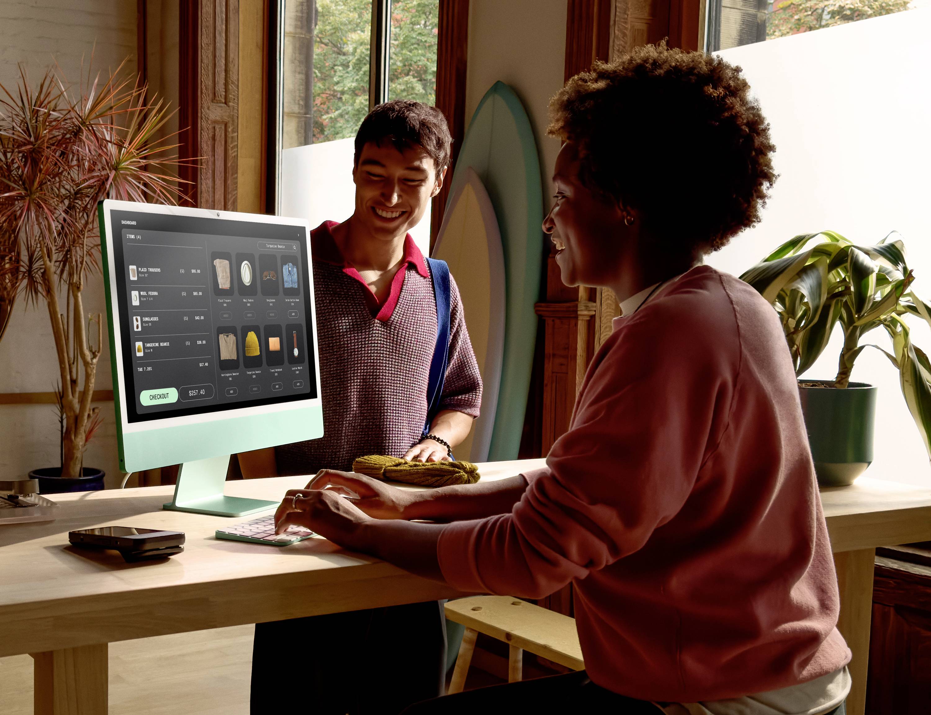 Zwei Personen interagieren in einem modernen Arbeitsbereich; eine Person arbeitet an einem Desktop-Computer mit geöffneter Design-Software. Große Fenster und Pflanzen sorgen für eine helle und freundliche Atmosphäre.