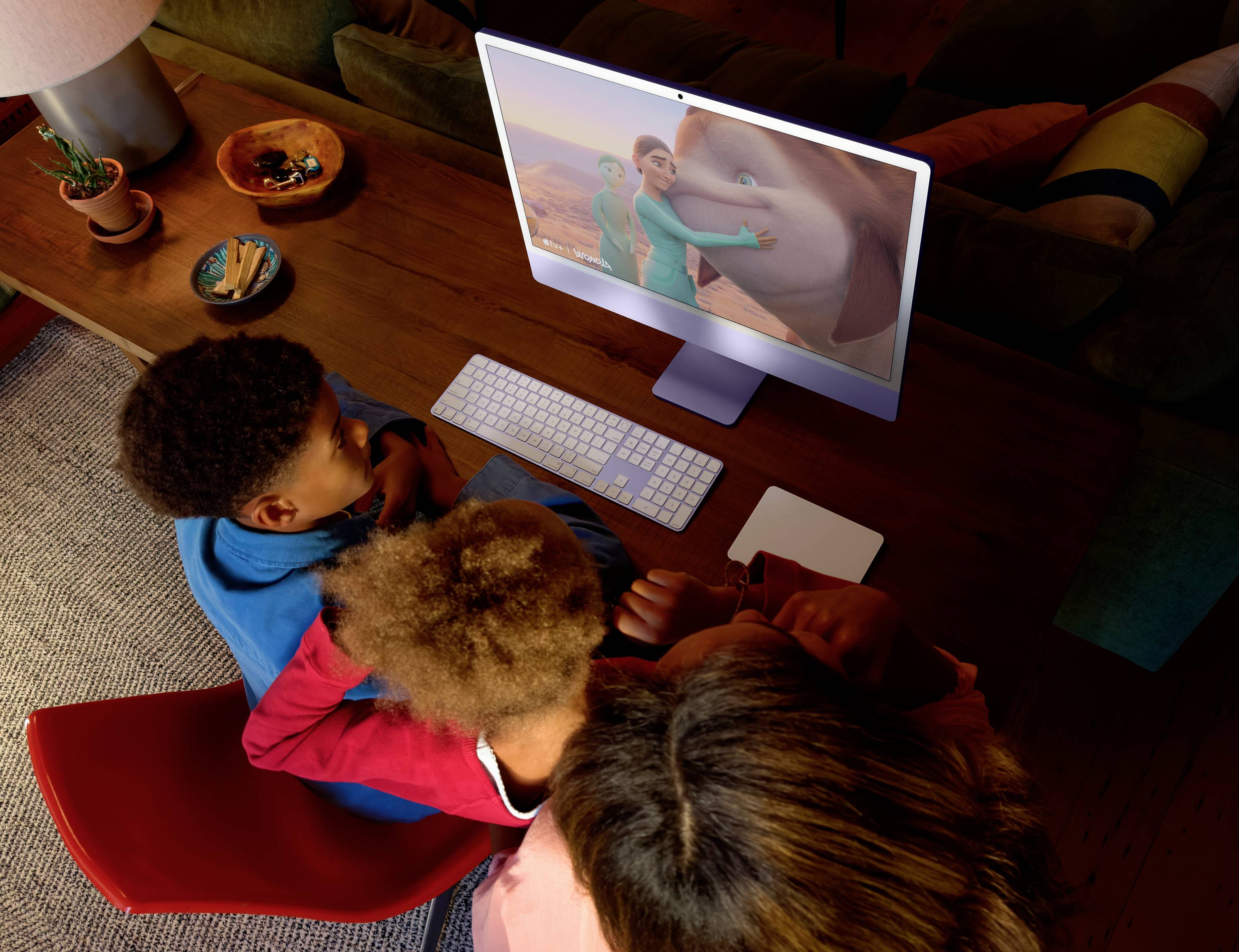 Eine Familie mit drei Personen schaut einen Animationsfilm auf einem Computerbildschirm in einem gemütlichen Wohnzimmer, umgeben von Kissen und Pflanzen.